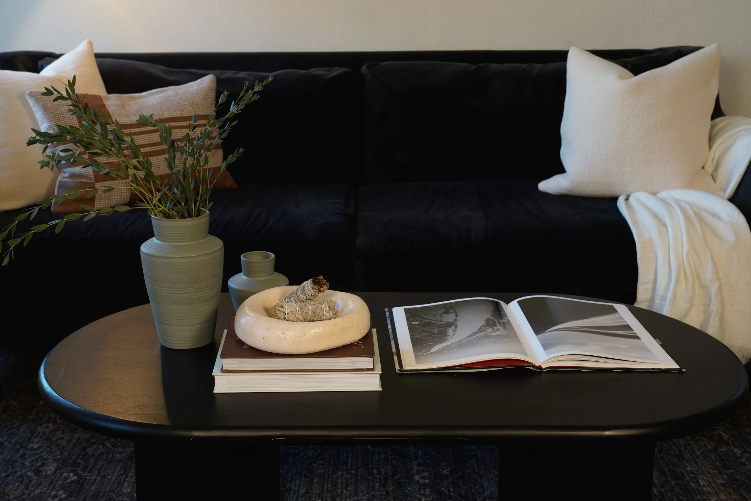 Living room with a dark sofa adorned with beige and white cushions, and a white blanket draped over the armrest. A black coffee table in front holds a large vase with green foliage, a small green vase, two stacked books, an open photo album, and a de