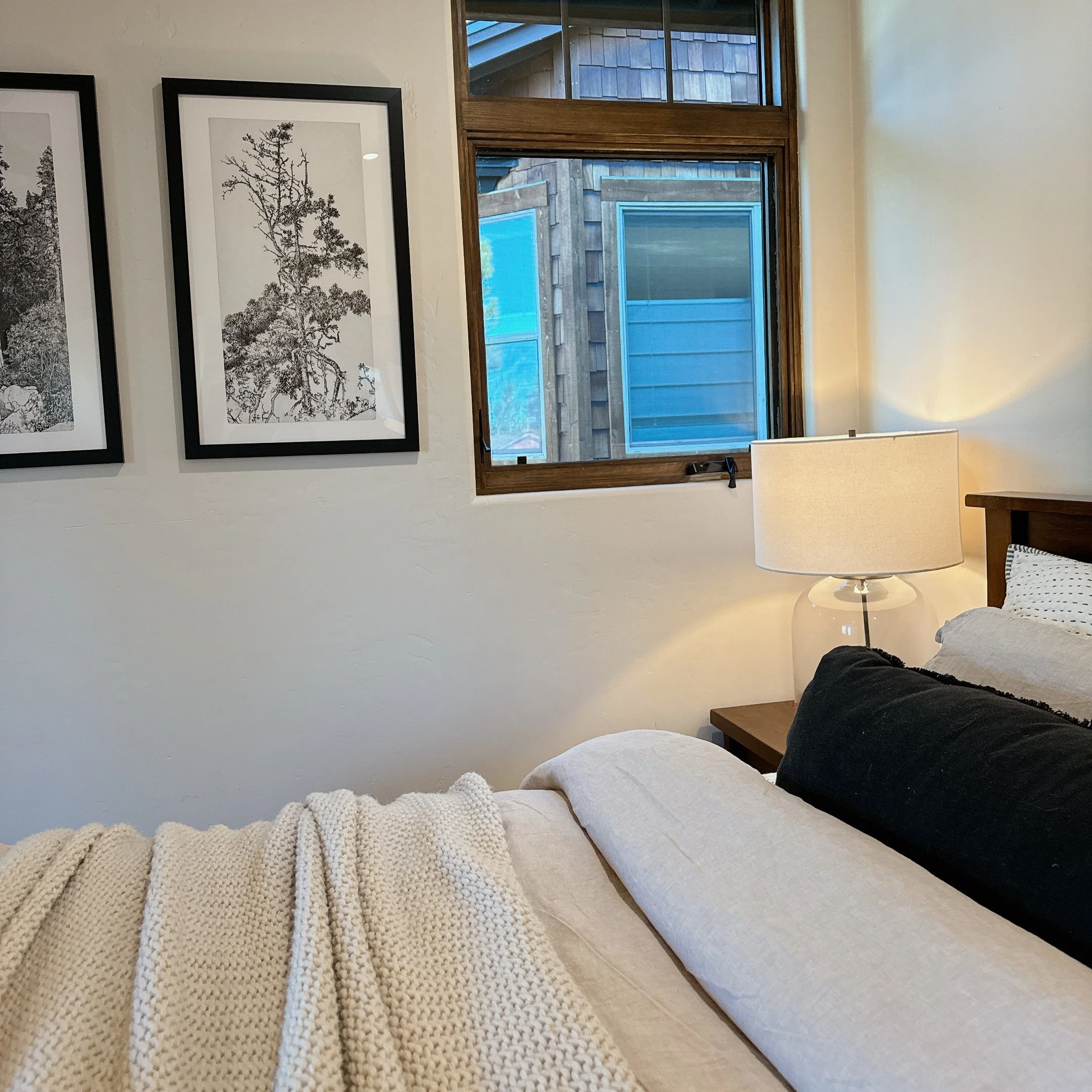 A cozy bedroom with framed black and white tree art on the wall, a bedside table with a white lamp, and a bed with white and black pillows and a knitted blanket. Perfect bed styling for home staging and every day living. 