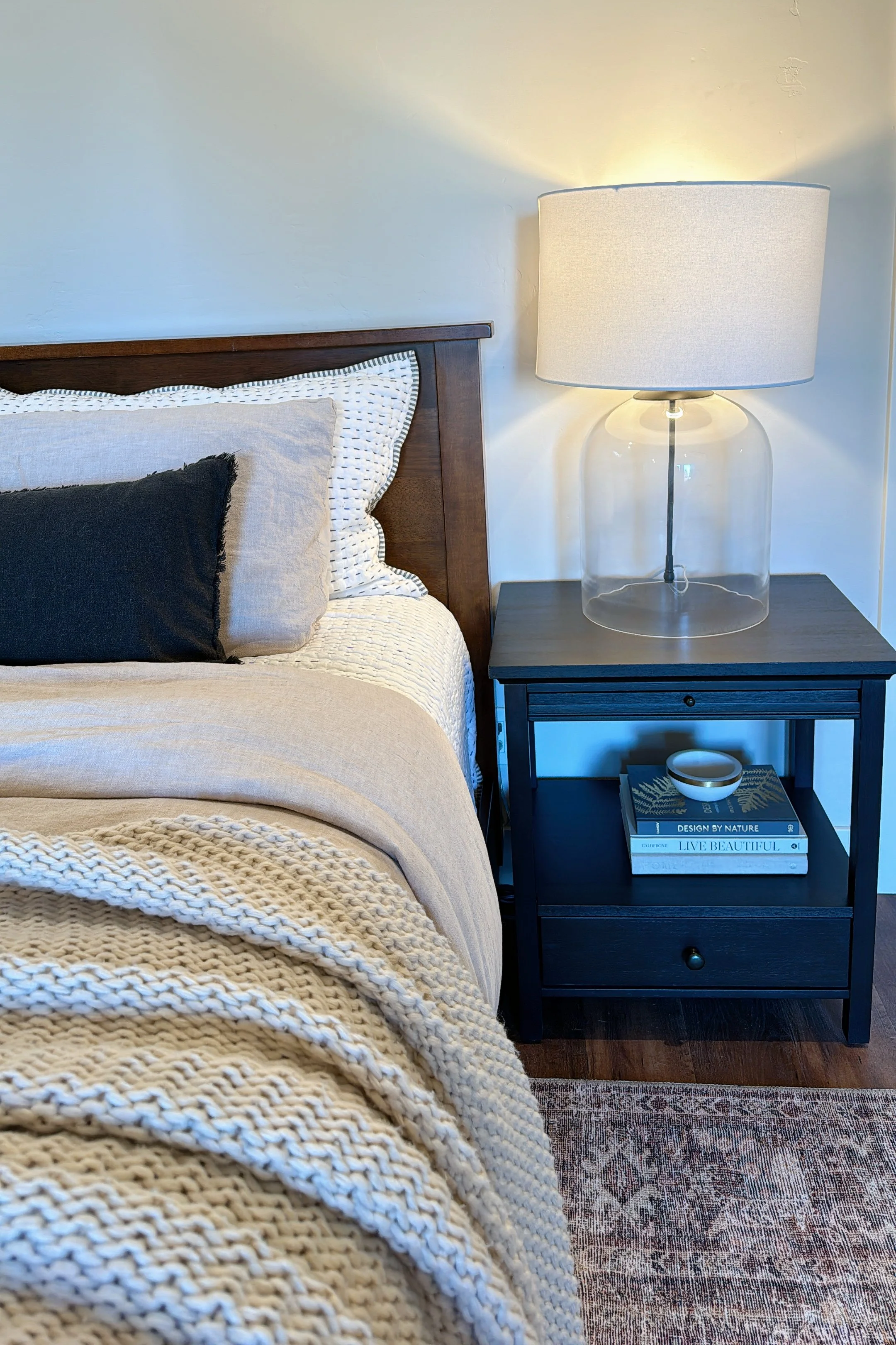 Warm, staged bedroom featuring a wood bed frame, layered neutral bedding, textured knit blanket, and styled nightstand with lamp and décor—demonstrating professional home staging and interior design.