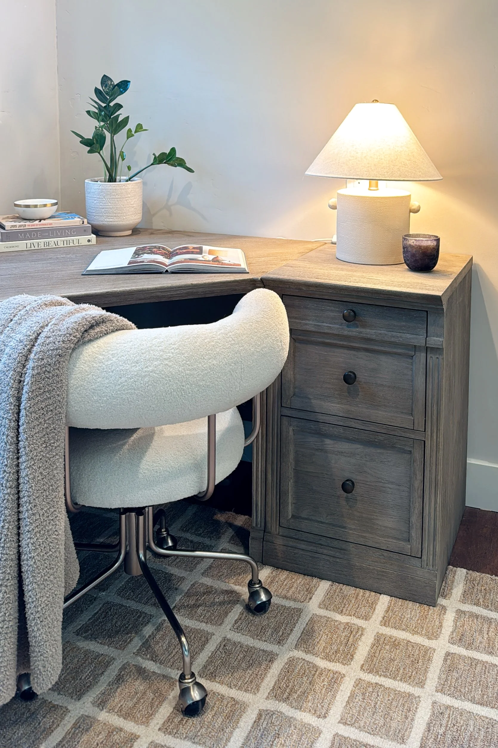 A cozy workspace corner with a wooden desk, a white upholstered office chair, a small table lamp, a potted plant, books, and a decorative bowl.