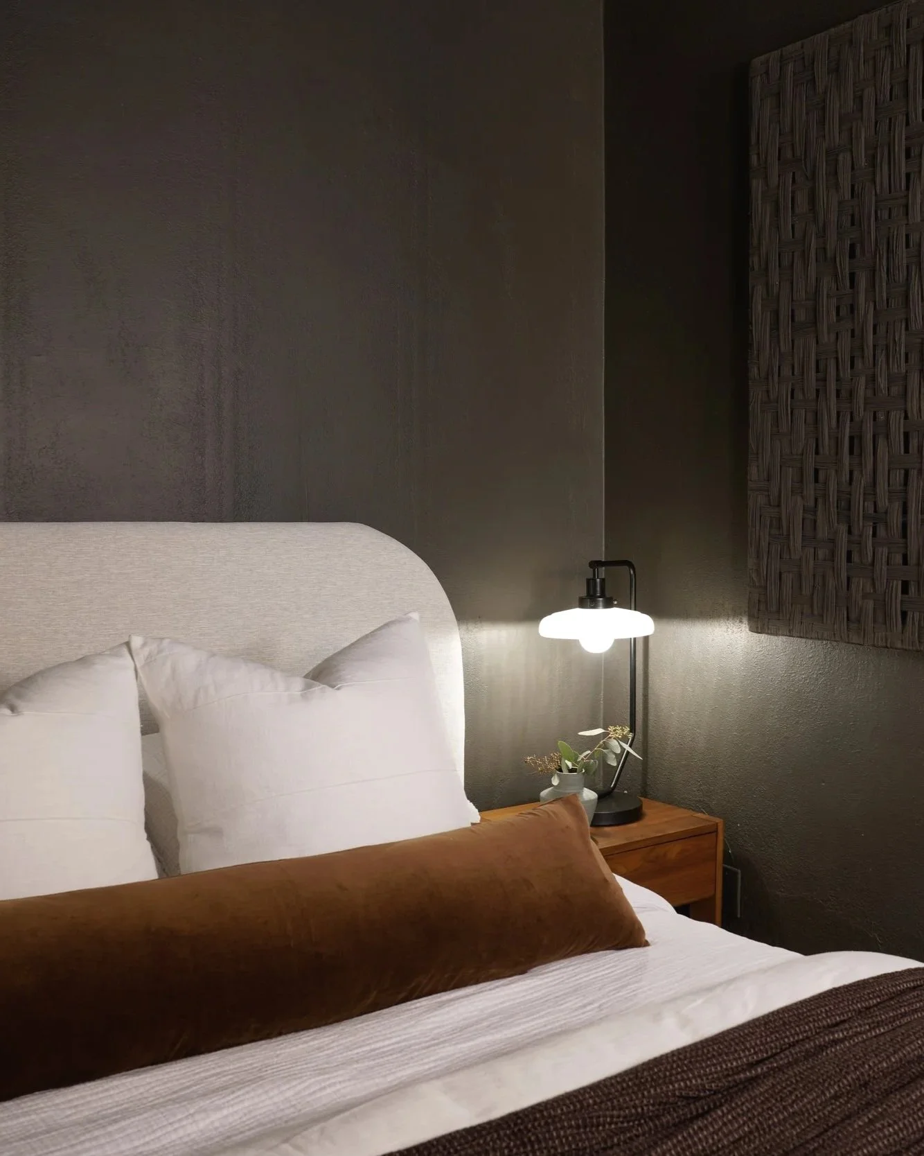 A cozy bedroom corner with a bed featuring white pillows and a brown bolster pillow. A wooden nightstand with a lamp and a small plant is beside the bed, against dark textured walls.