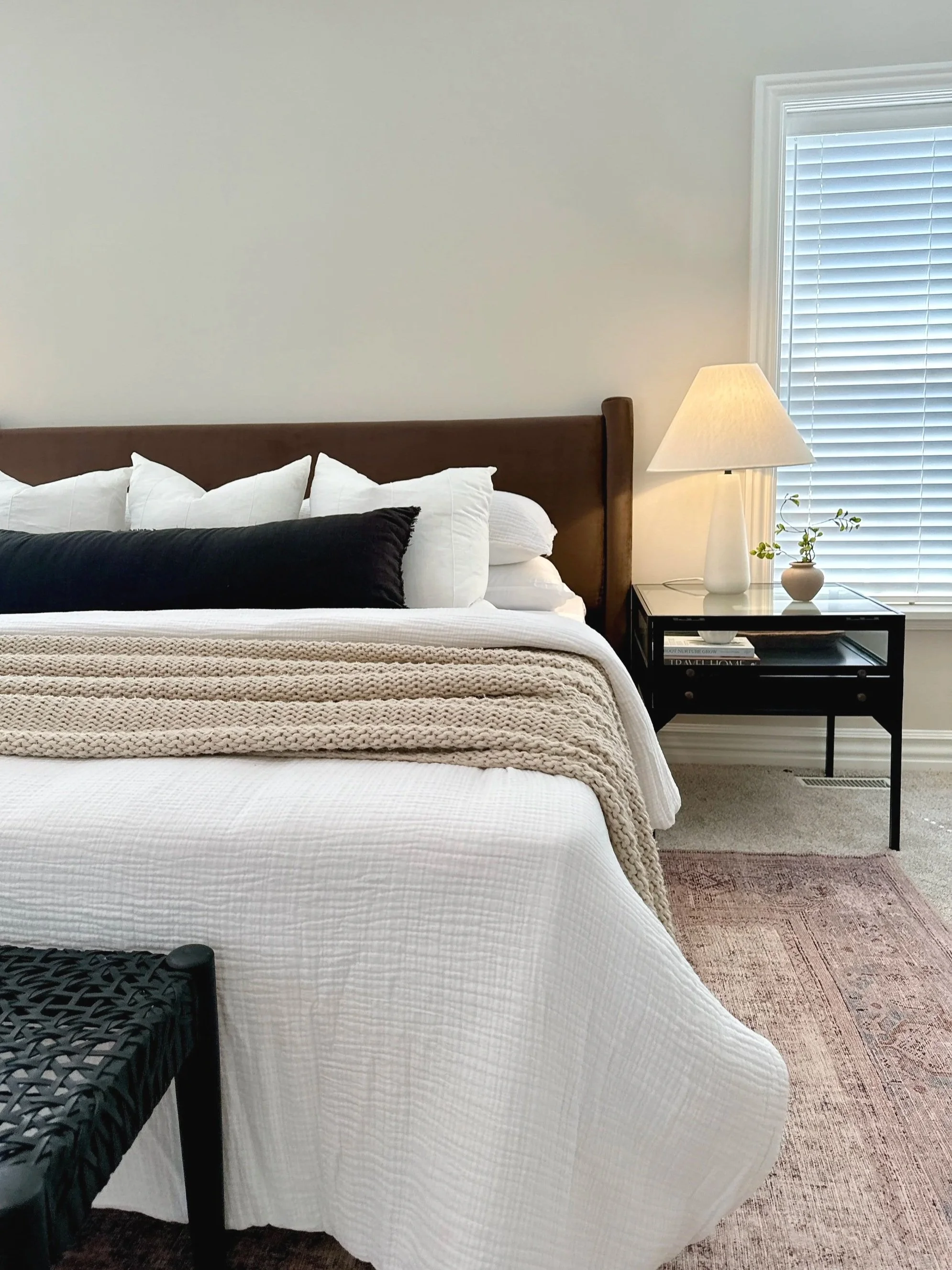 Staging by Riley James Interiors in Utah. A bedroom with a white bedspread, multiple pillows, a woven blanket, a black side table with books and a lamp, a window with blinds, and a beige rug.