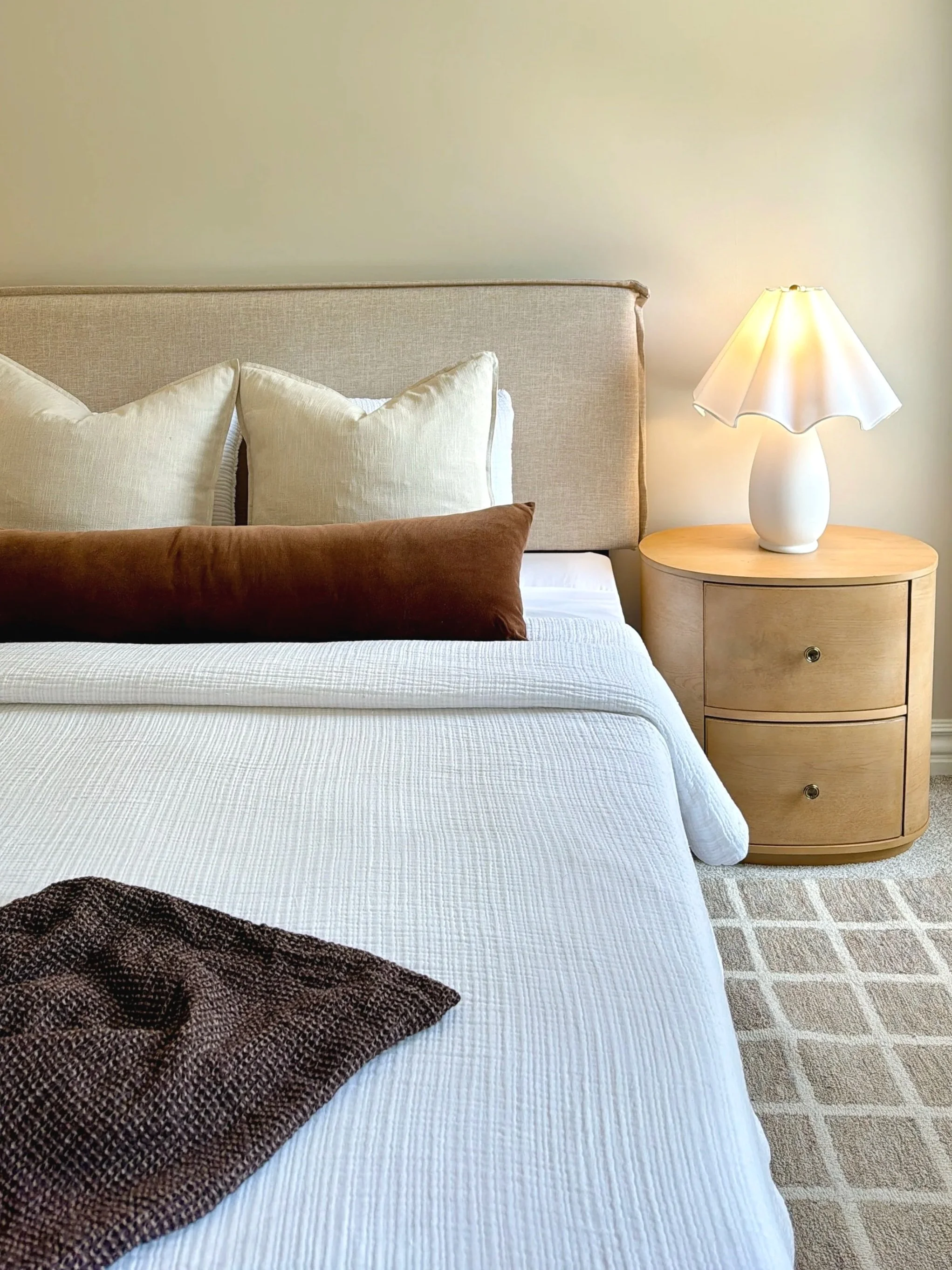 Bedroom with beige upholstered headboard, white bedding, tan and brown pillows, a beige nightstand with two drawers, a white table lamp with a scalloped shade, and a patterned beige carpet. Staging by Riley James Interiors.