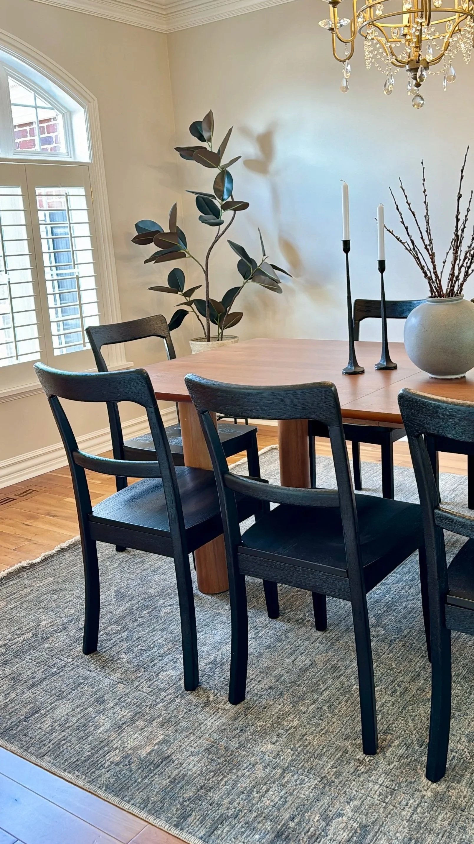Dining room with a wooden table, black chairs, a large window with shutters, a chandelier, a potted plant, candlesticks, and a vase with branches. Vacant and occupied staging services in Salt Lake City, Utah by Riley James Interiors.