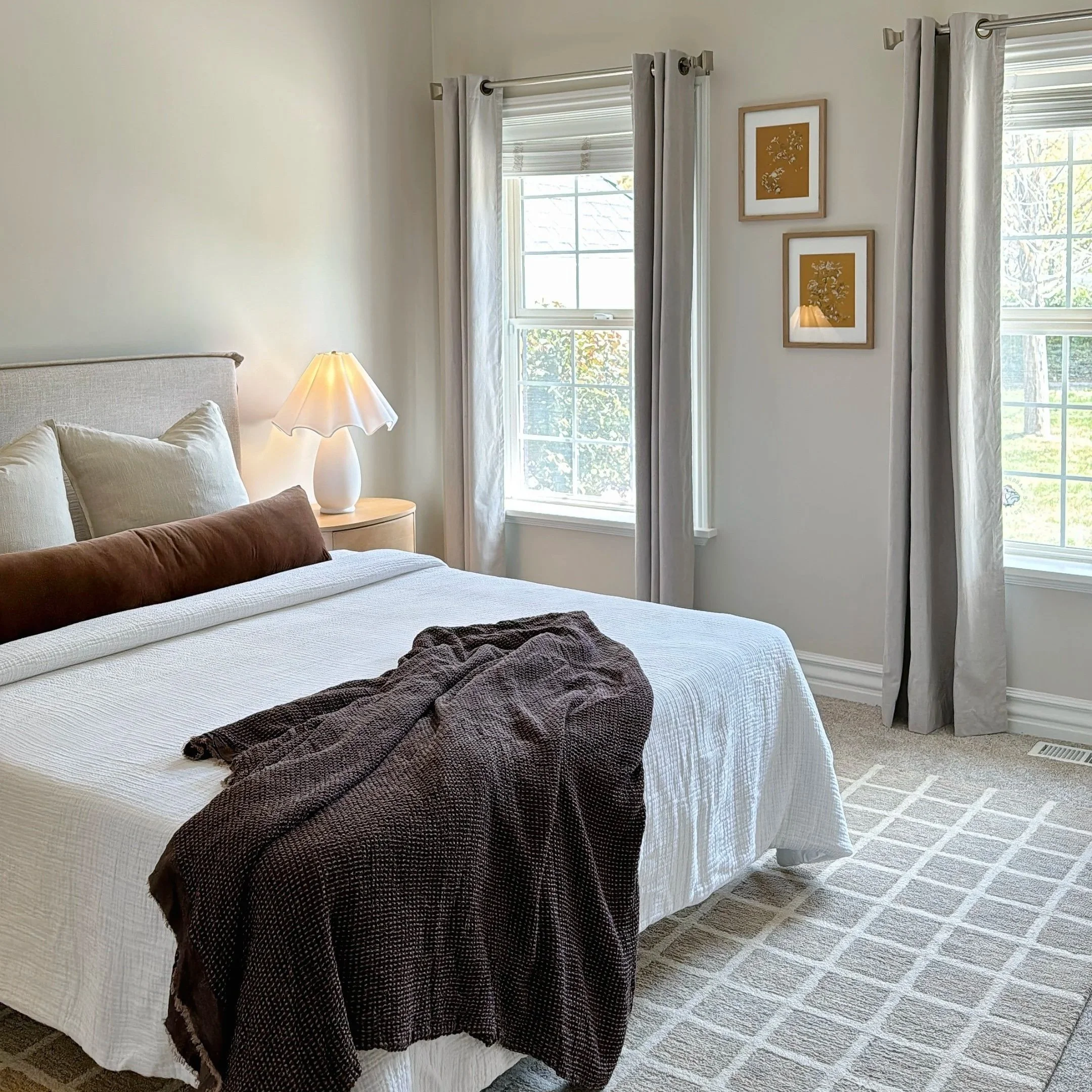 Home staging and interior design by Riley James Interiors in Salt Lake City, Utah. A bedroom with a bed, two windows with curtains, a bedside lamp, and framed wall art. Natural light is coming through the windows.