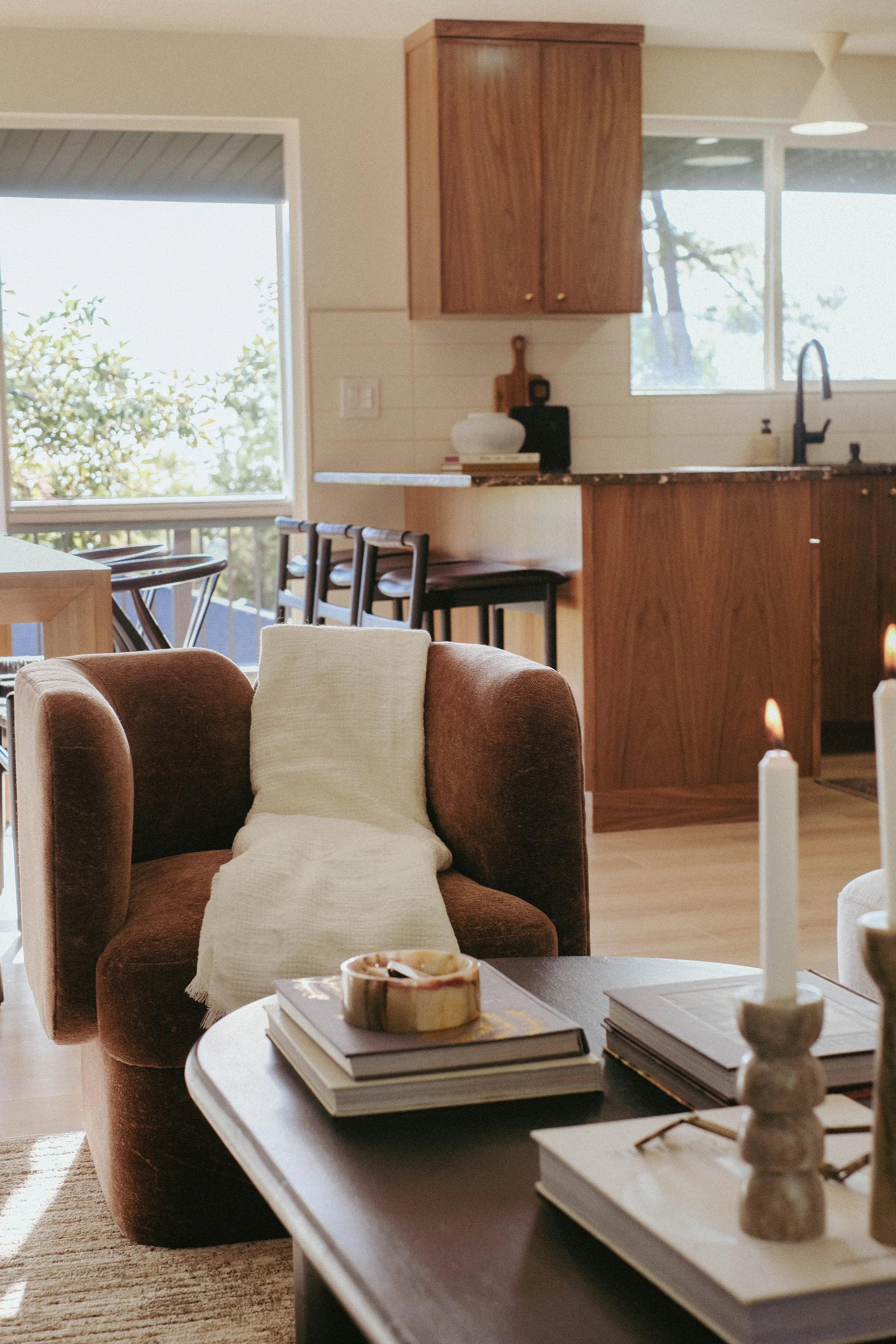 Living room with brown upholstered armchair, cream cushion, coffee table with books, and decorative objects, open kitchen with wooden cabinets and bar stools, large windows with outdoor view
