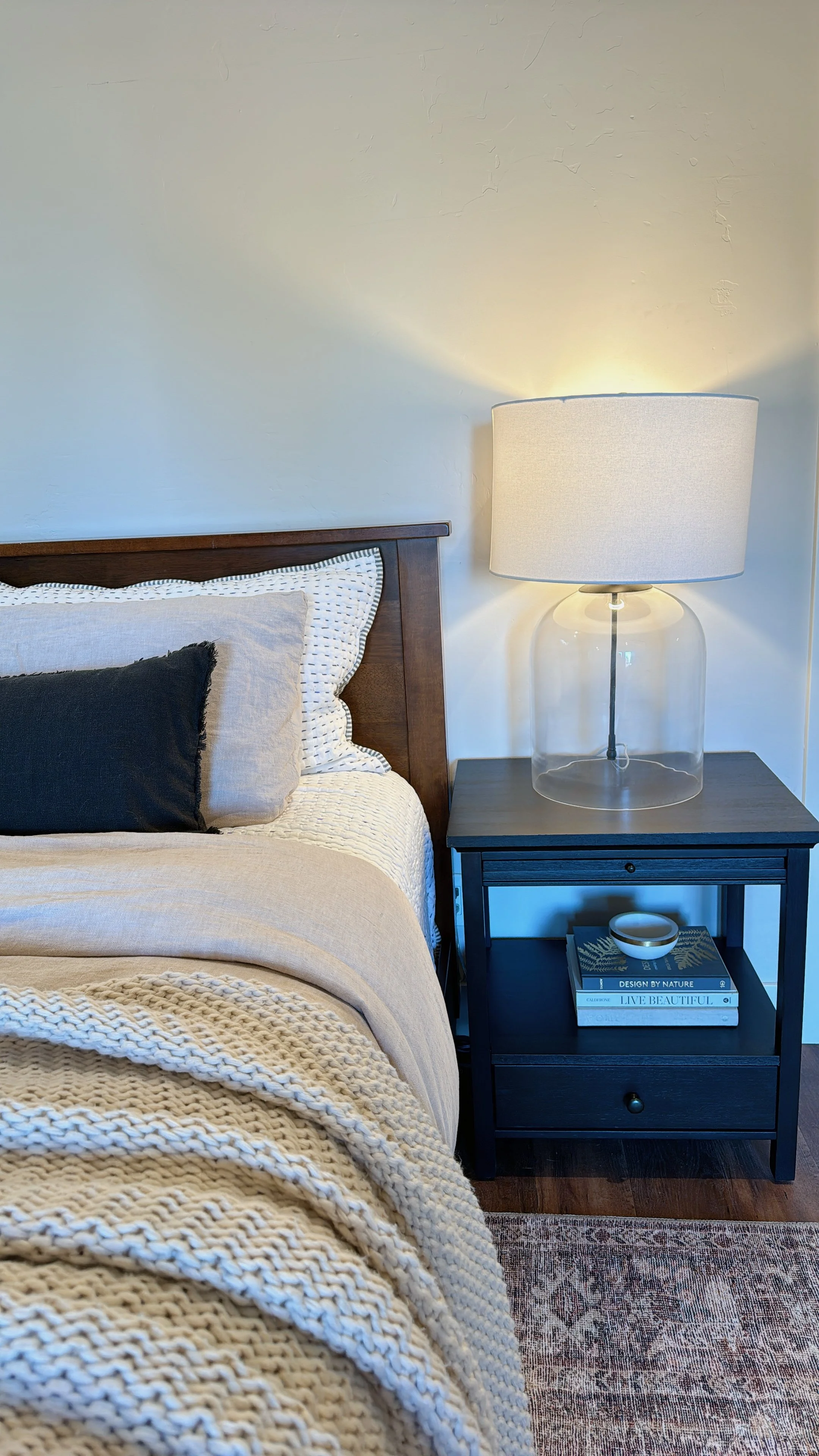 Close-up of a bedroom corner with a wooden bed frame, beige and black pillows, a textured beige blanket, a black nightstand with books, a small bowl, and a modern glass lamp, next to a white wall and a patterned rug.