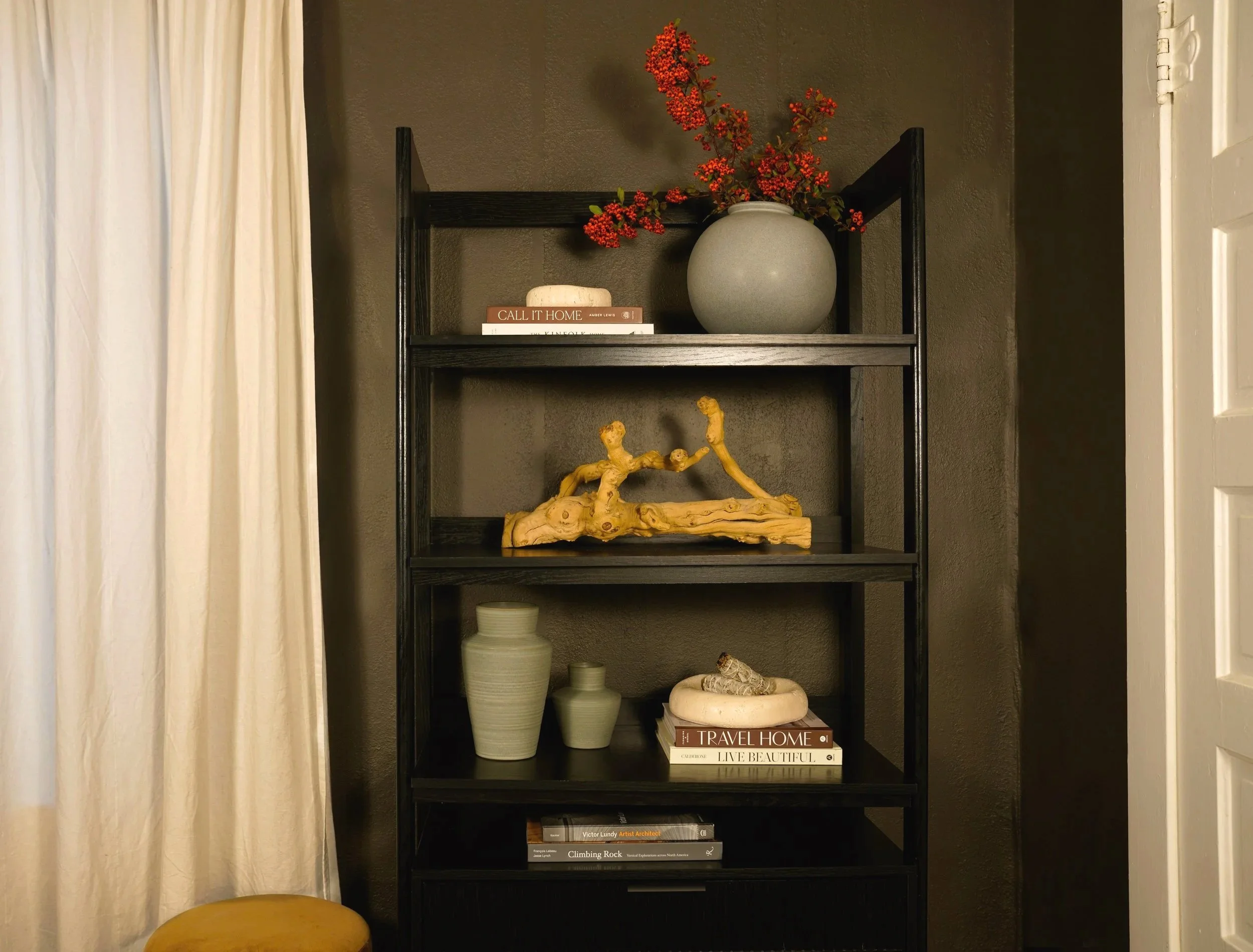 A black bookshelf with four shelves. The top shelf has a round gray vase with red berries and a stack of three books. The second shelf has a piece of driftwood and a small figurine. The third shelf has two light green vases and a white dish with decorative items, along with two stacked books. The bottom shelf has a few books. To the left is a cream-colored curtain, and to the bottom left corner there is a yellow cushion. The background wall is dark gray.