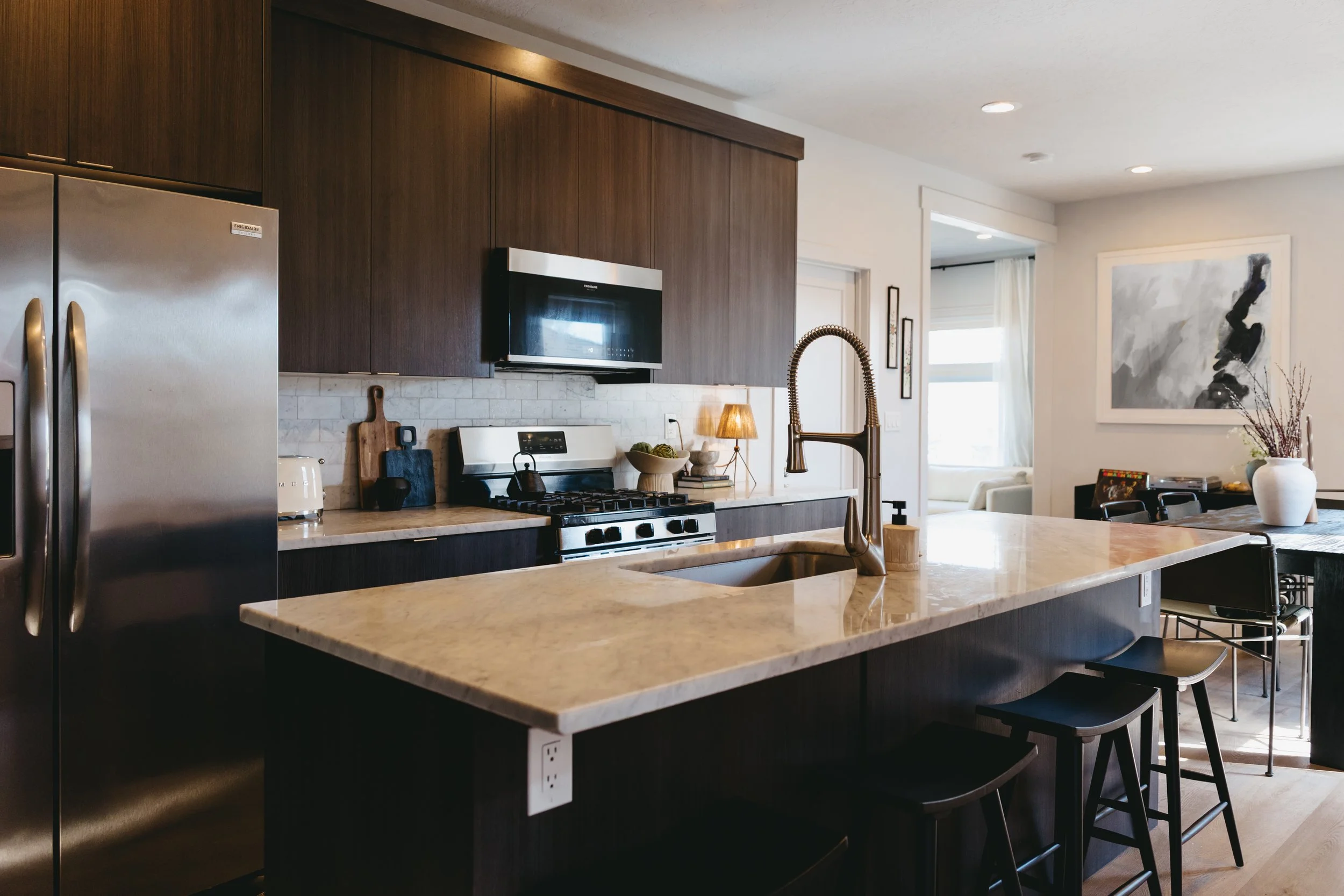 Modern kitchen with dark wood cabinets, a marble island with a sink, stainless steel appliances, including a refrigerator and oven, and decorative elements like a bowl of fruit, a small lamp, and framed artwork on the wall. Riley James Interiors.