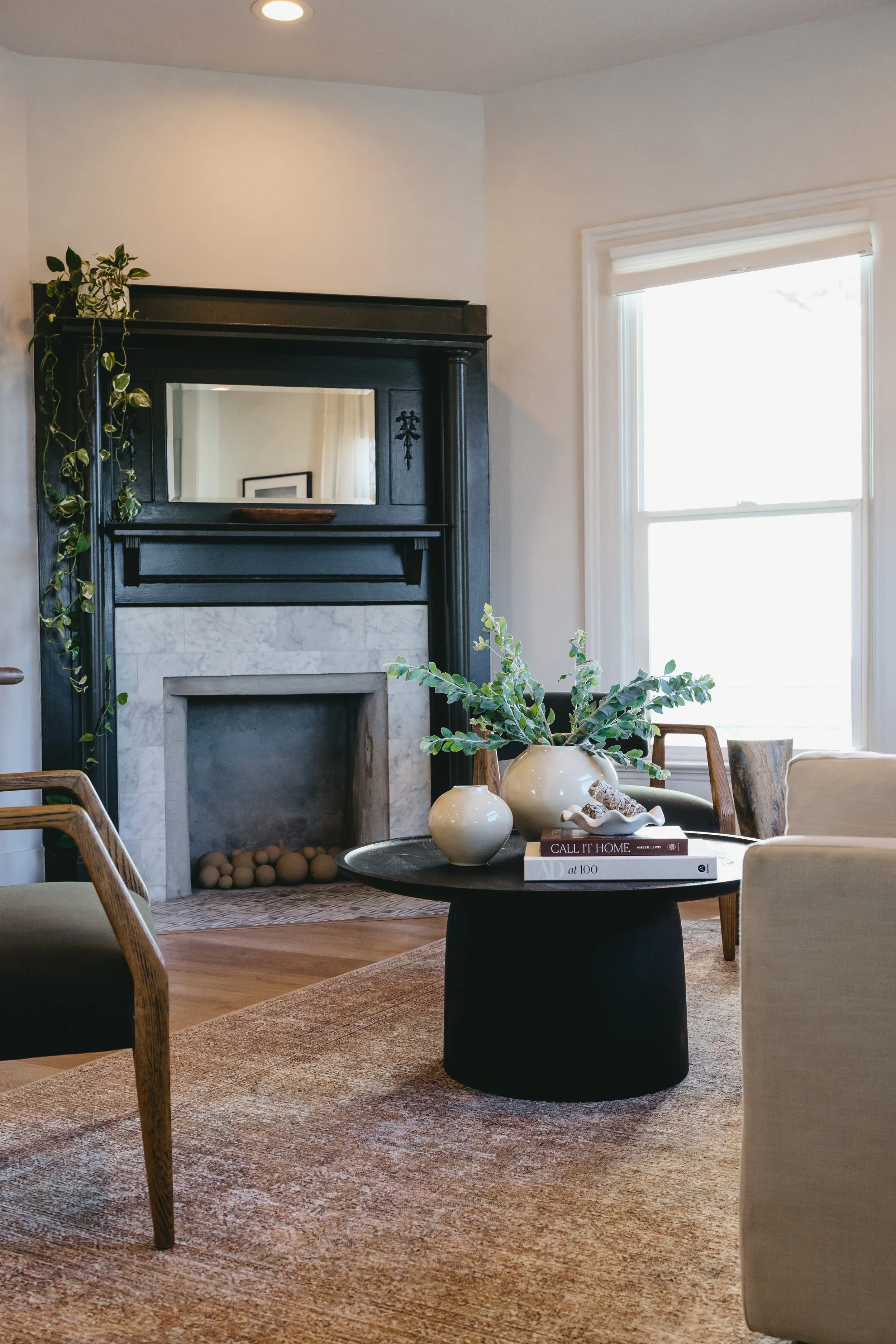 Vacant stage by Riley James Interiors in Utah. Living room with a black mantel fireplace, a round black coffee table with decorative vases and books, beige armchair, wooden chairs, a window with white blinds, and a beige rug.