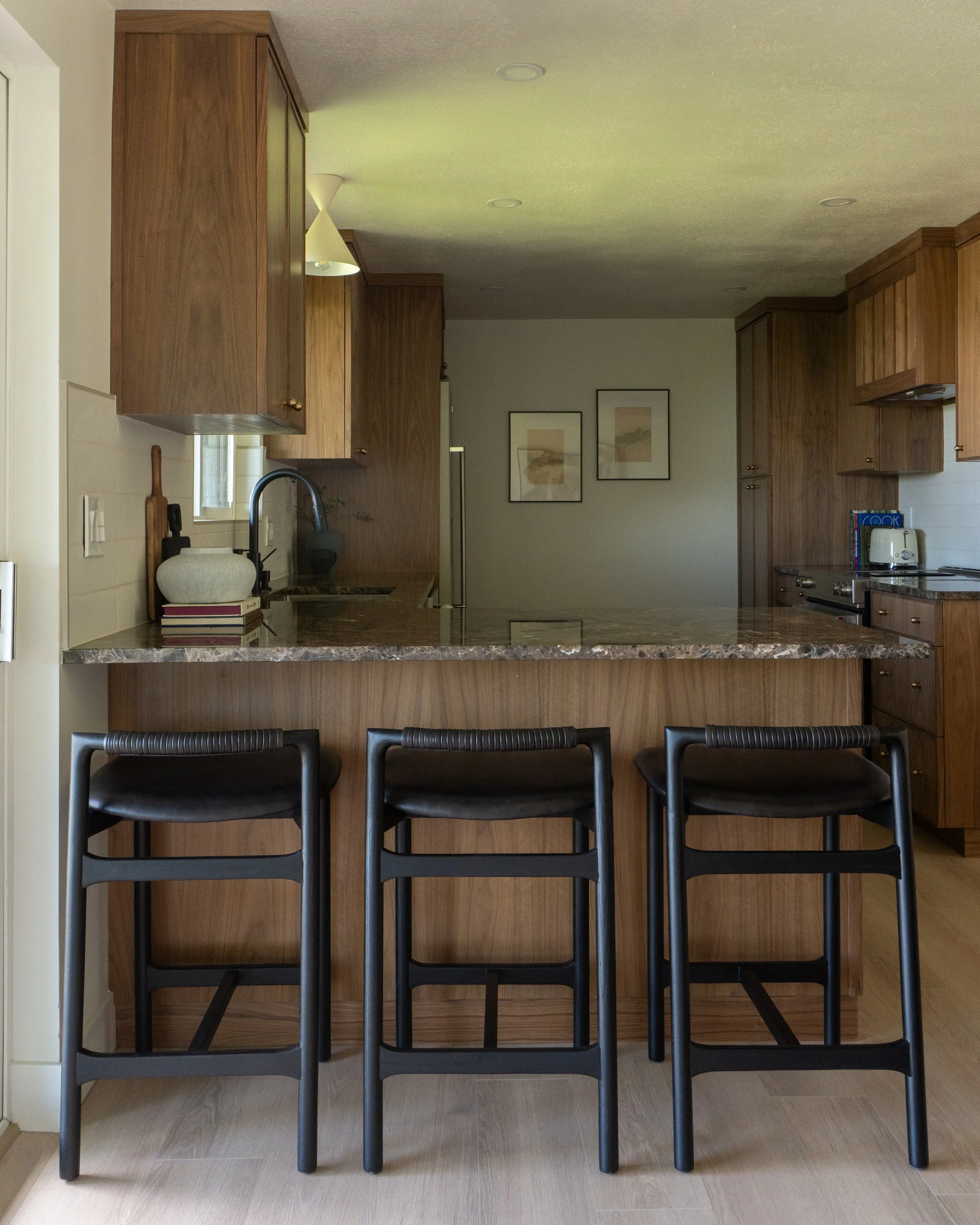 Kitchen with black bar stools, wooden cabinets, beige tiled floor, and a marble countertop. Three pictures on the wall are visible, along with a countertop appliance.