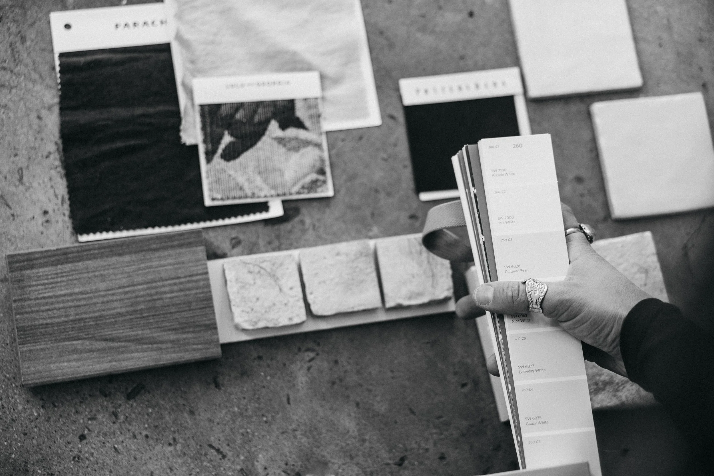 A person holding a paint color sample fan deck next to a variety of paint swatches, tile samples, and fabric swatches laid out on a surface, likely for interior design or renovation planning.