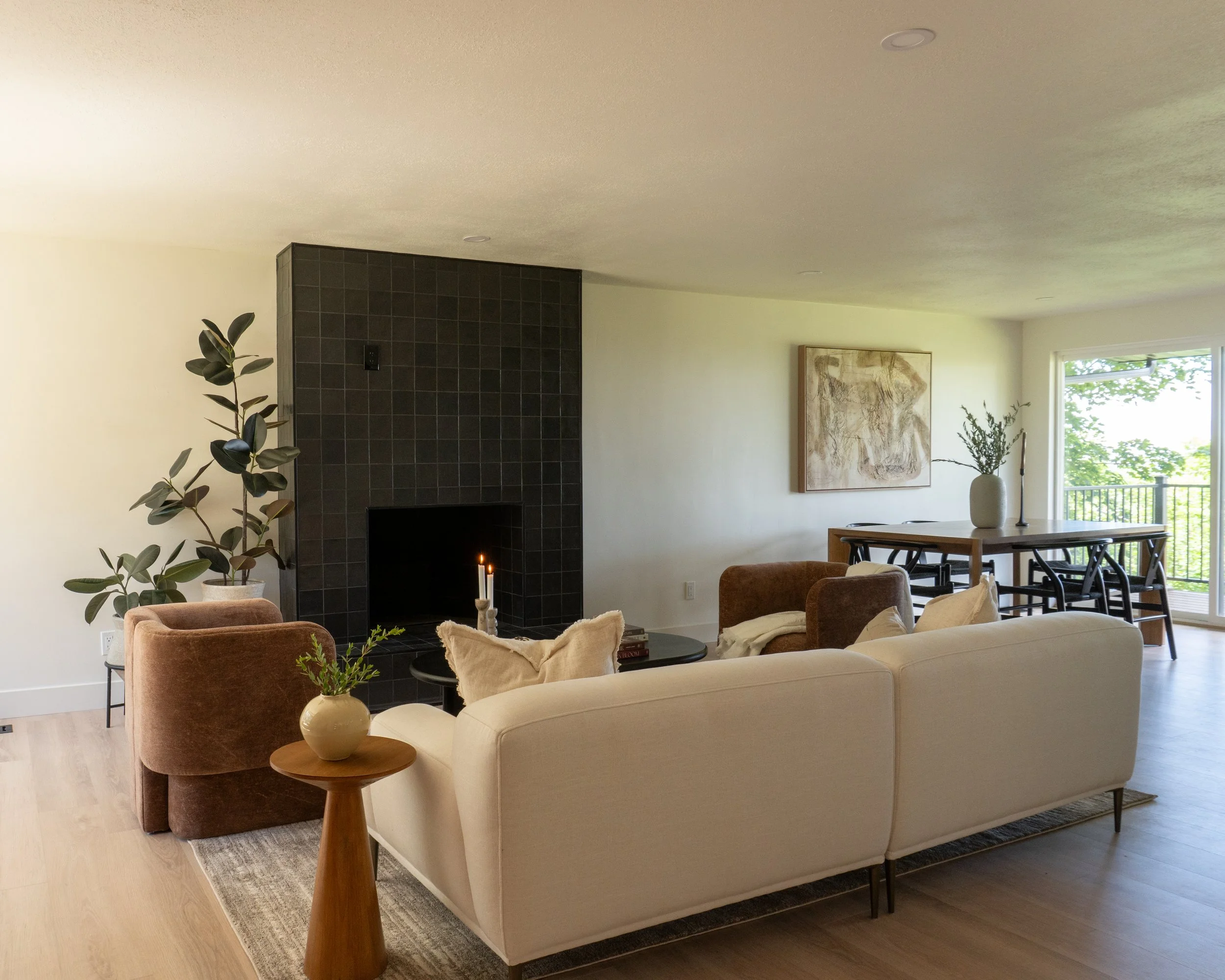 Living room with beige sofa, dark armchair, coffee table with a vase and decor, fireplace with candles, framed artwork, window with outdoor trees, bookshelf, and plants.