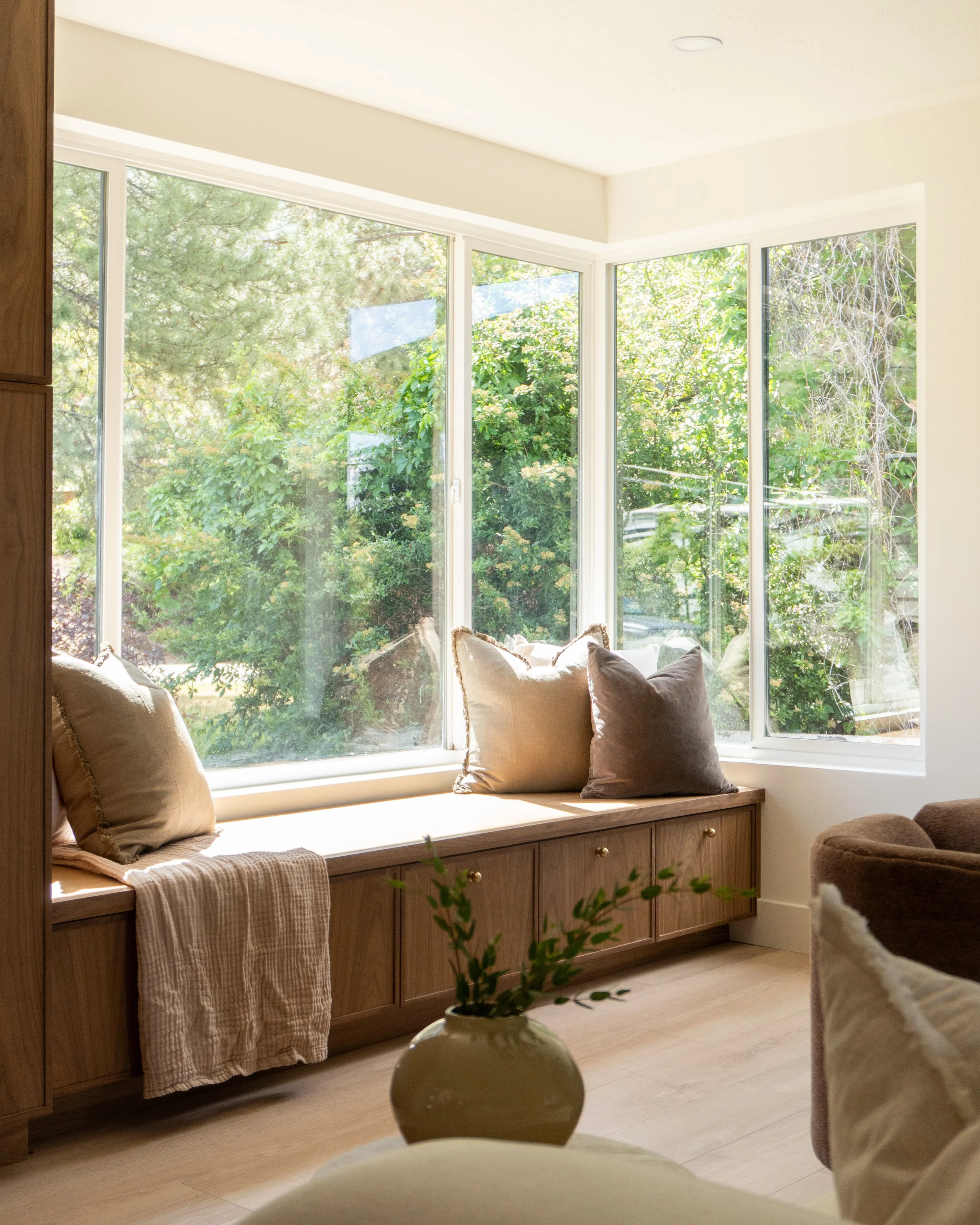 Living room window seat with beige cushions and throw blanket, large windows showing green trees outside, potted plant on table in foreground.