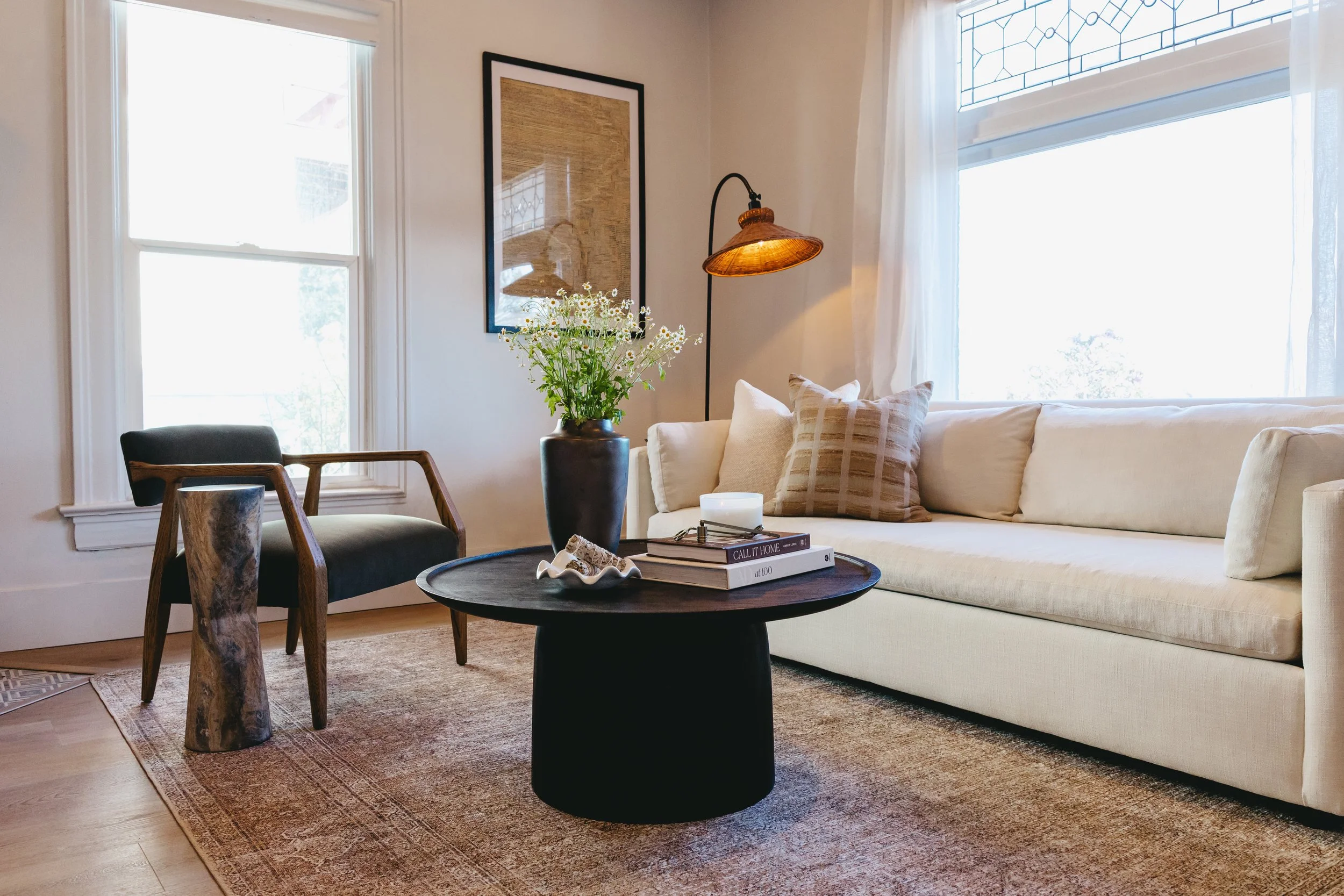 Staged living room with cream sofa, window with white curtains, wood and black accent chair, black round coffee table with books and vase of flowers, wall art, standing lamp, area rug by Riley James Interiors.