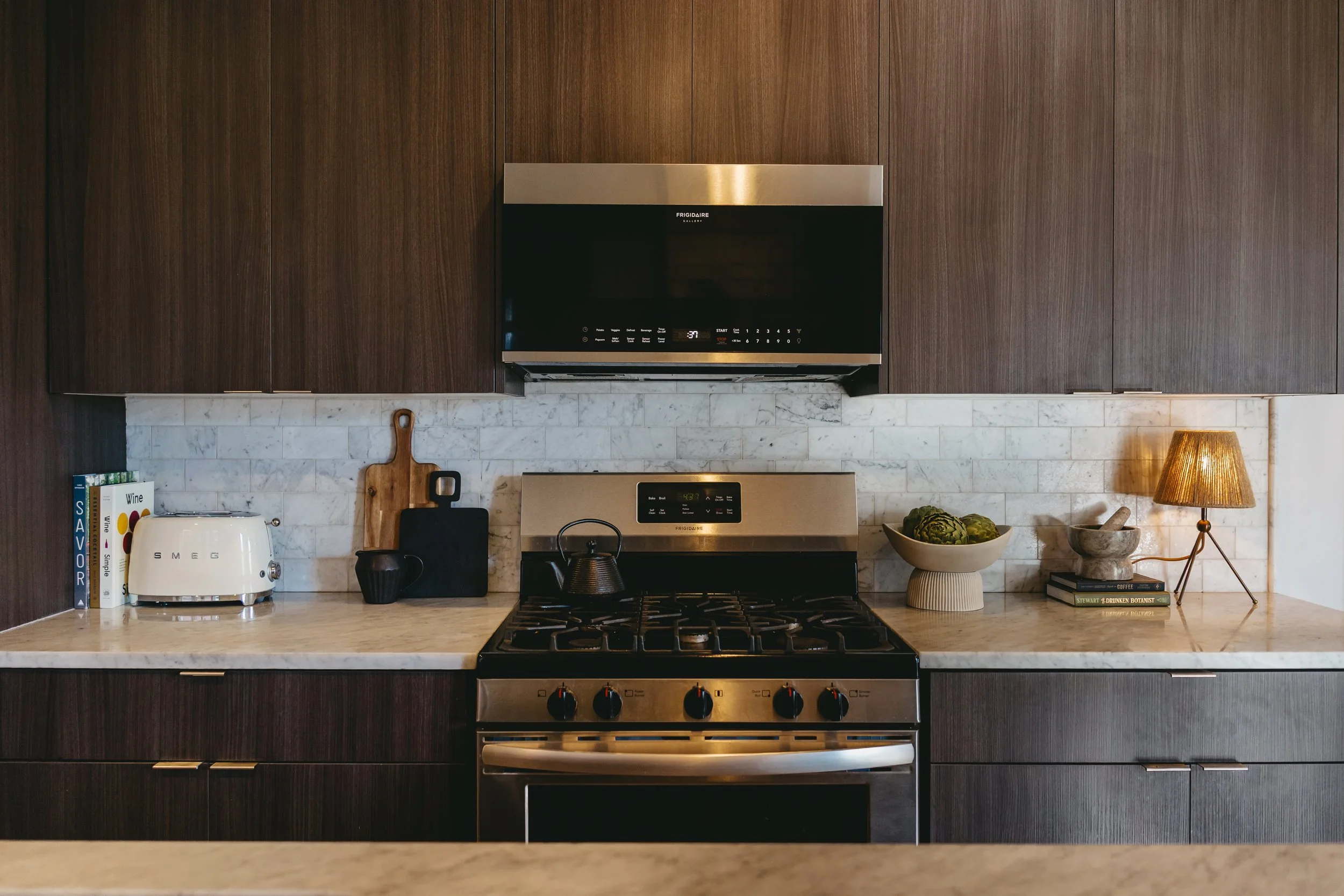 Modern kitchen with dark wood cabinets, a marble backsplash, stainless steel stove and microwave, a white toaster, cutting boards, a bowl of artichokes, a mortar and pestle, books, and a small table lamp. Staging by Riley James Interiors in Utah.