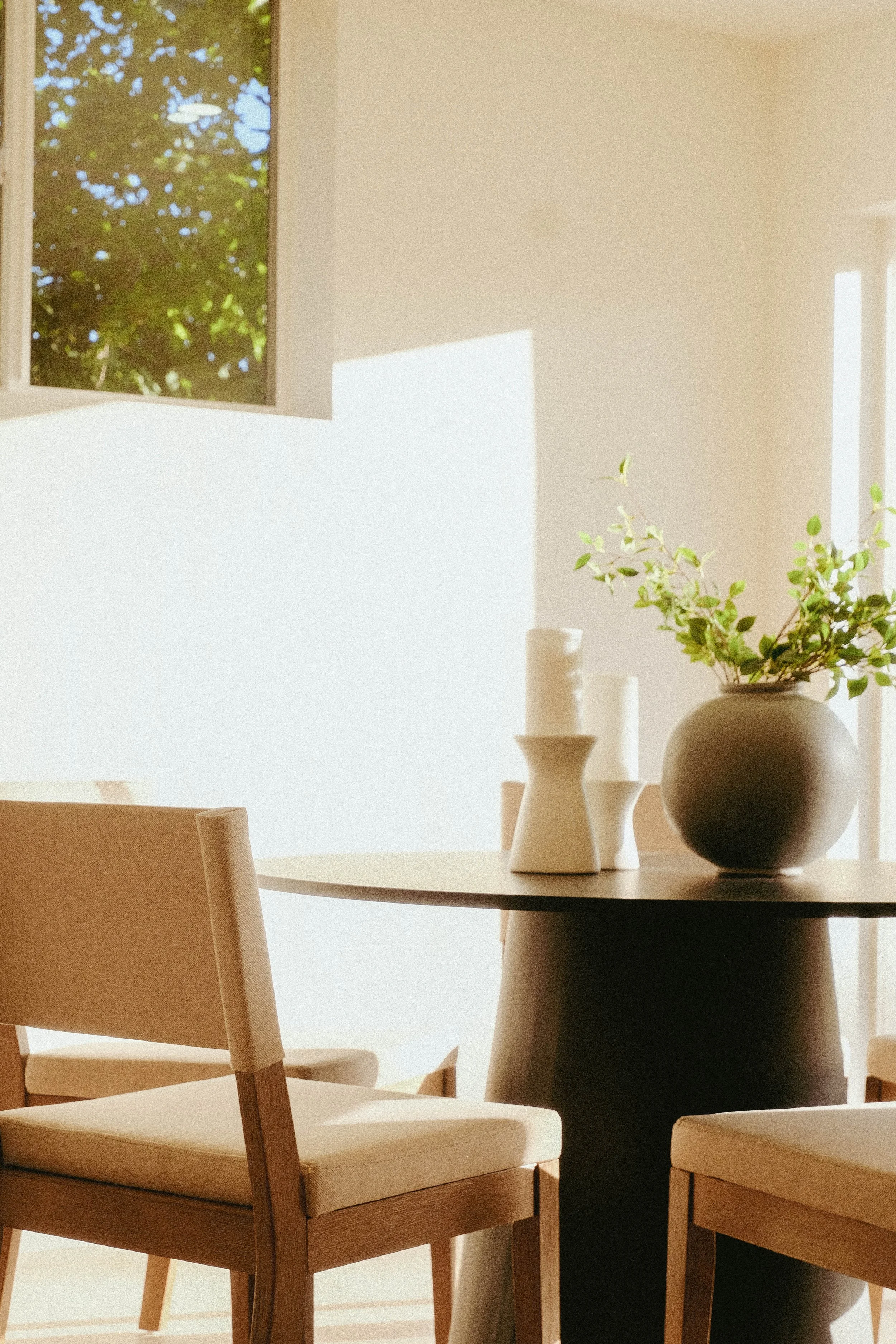 A dining room with a round table, beige chairs, and a large black vase with green leafy branches. There are white candles on the table. A window shows green foliage outside and sunlight streams into the room.
