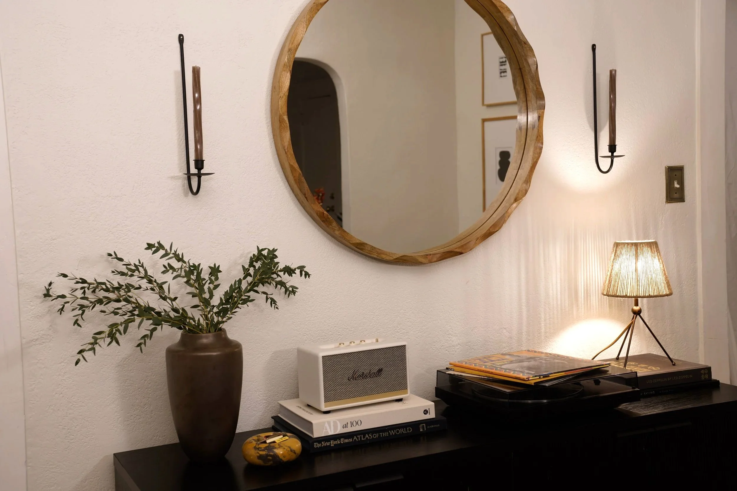 A living room wall decorated with a round wooden-framed mirror flanked by black candle wall sconces, a black sideboard holding a brown vase with green leafy branches, a white Marshall speaker, a stack of books, a decorative rock, and a small table lamp with a beige shade.
