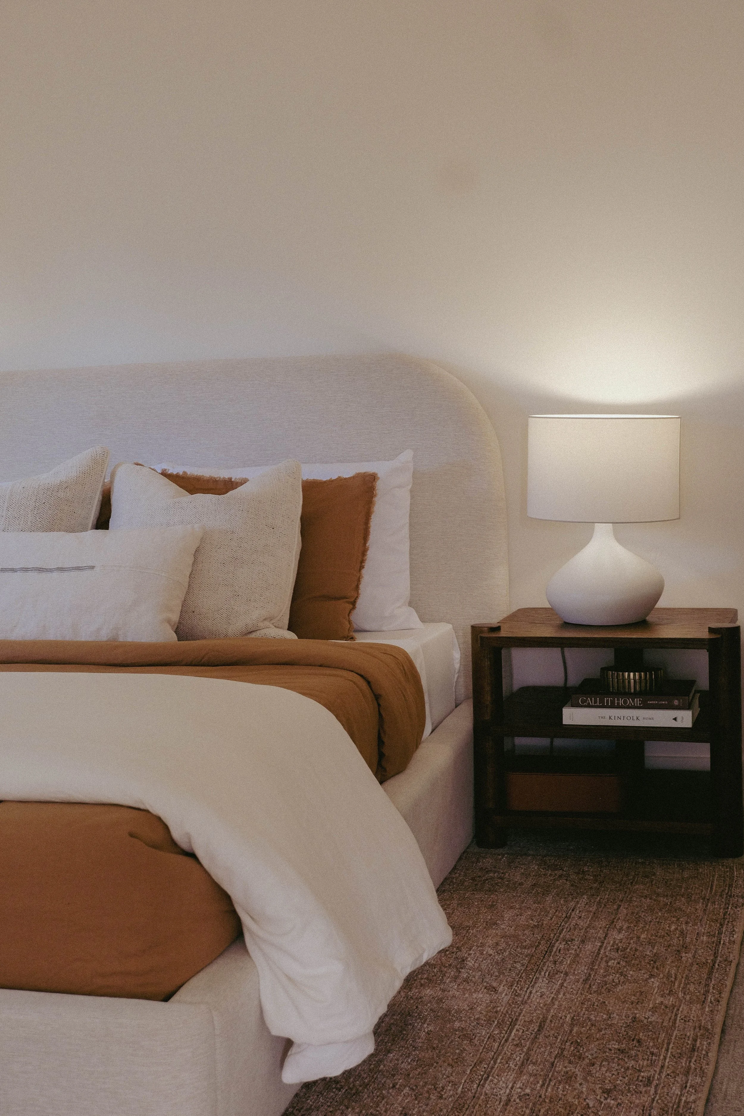 A cozy bedroom with a bed featuring beige and white pillows, a warm brown blanket, and a wooden nightstand holding a white lamp with a textured base and a stack of books, including titles about home and interior design.