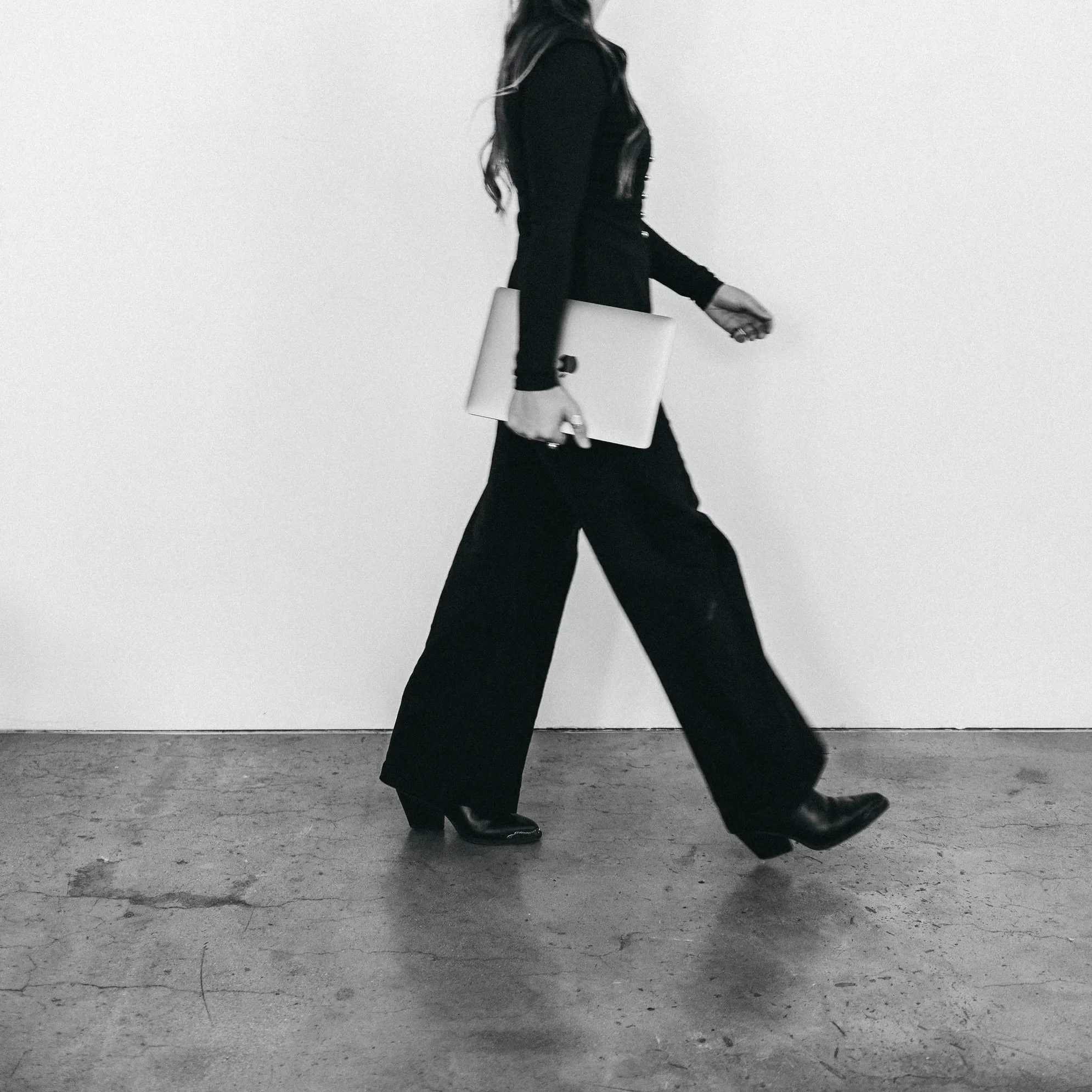 A woman in black clothing, holding a laptop in her left hand and a smartphone in her right hand, walking on a concrete floor against a plain white wall.