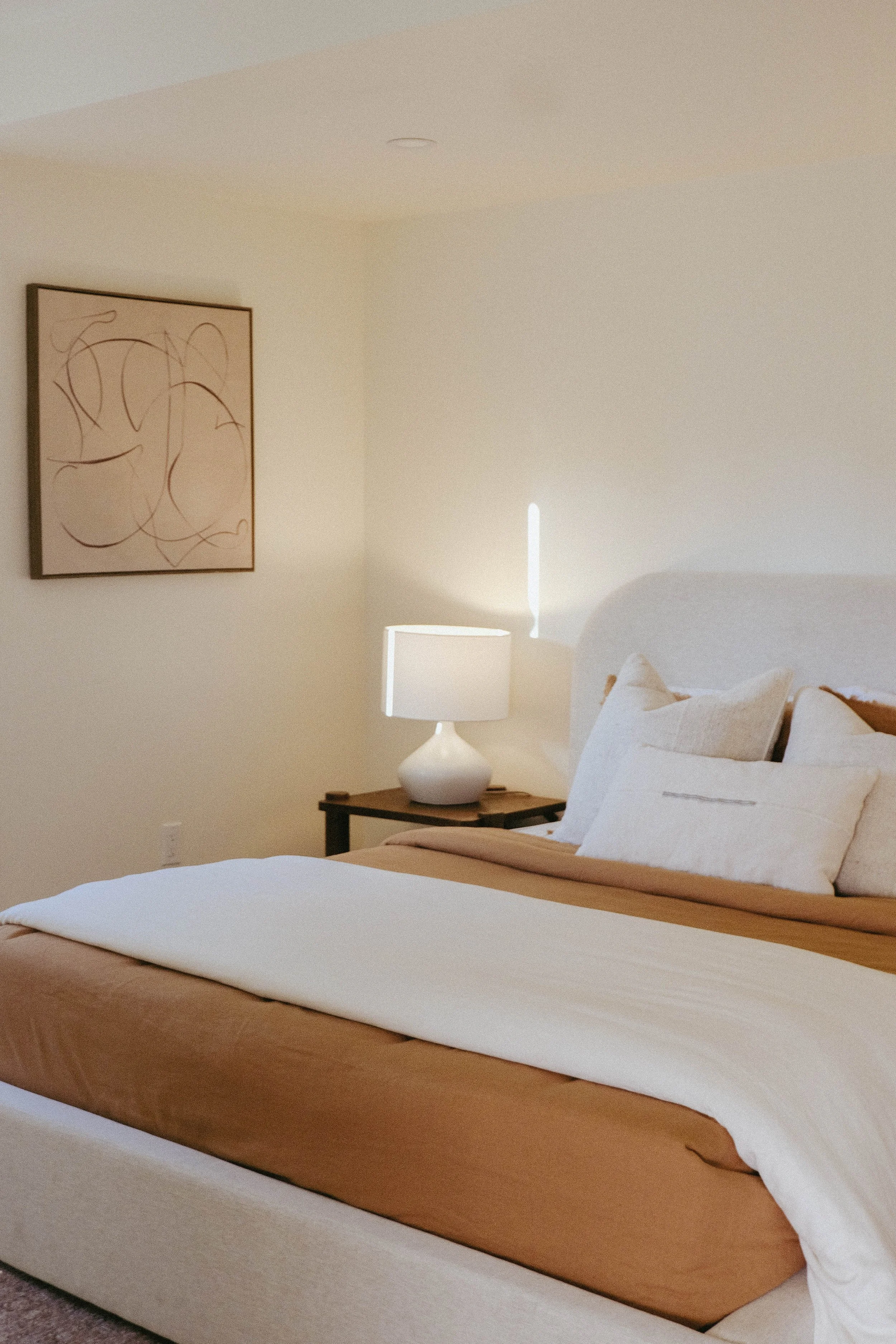 A neatly made bed with white sheets, beige blanket, and beige pillows in a minimalistic bedroom. A white table lamp sits on a small wooden nightstand beside the bed. A framed abstract artwork hangs on the cream-colored wall.
