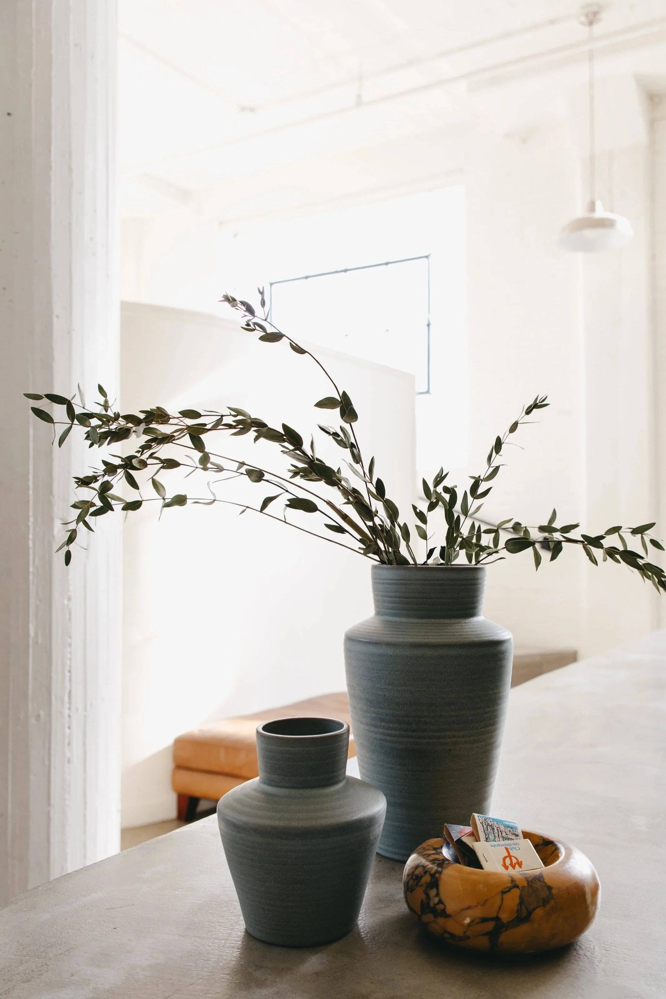 Interior scene with two gray vases, one with leafy green branches, placed on a table. A small yellow and brown marble bowl with cards inside is also on the table. Bright natural light fills the room with a white wall and window in the background.