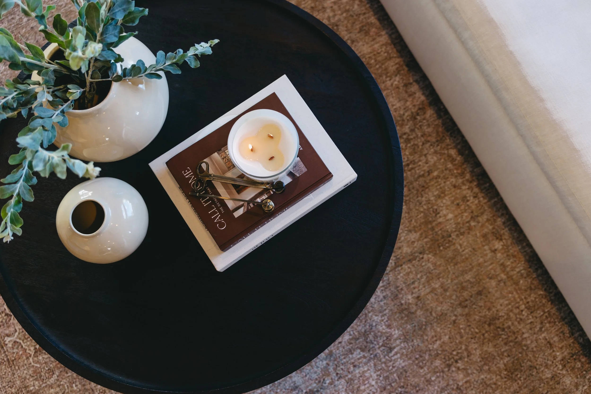 A black round coffee table holding a white ceramic vase with green foliage, a small round white ceramic vase, a stack of books with a white candles on top, and a pair of metal scissors. Interior styling services by Riley James Interiors in Utah.