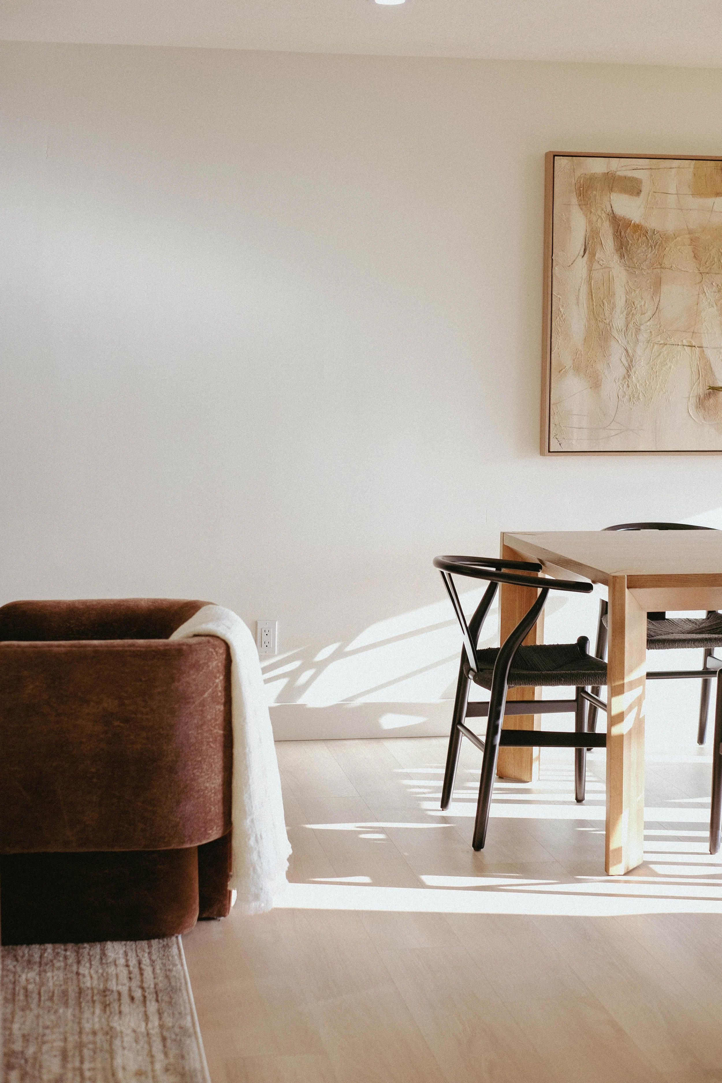Light-colored living room with a brown armchair on the left, a beige area rug, and a wooden dining table with two black chairs near a plain white wall. There is a large abstract painting with earth tones hanging on the wall. Sunlight casts shadows on