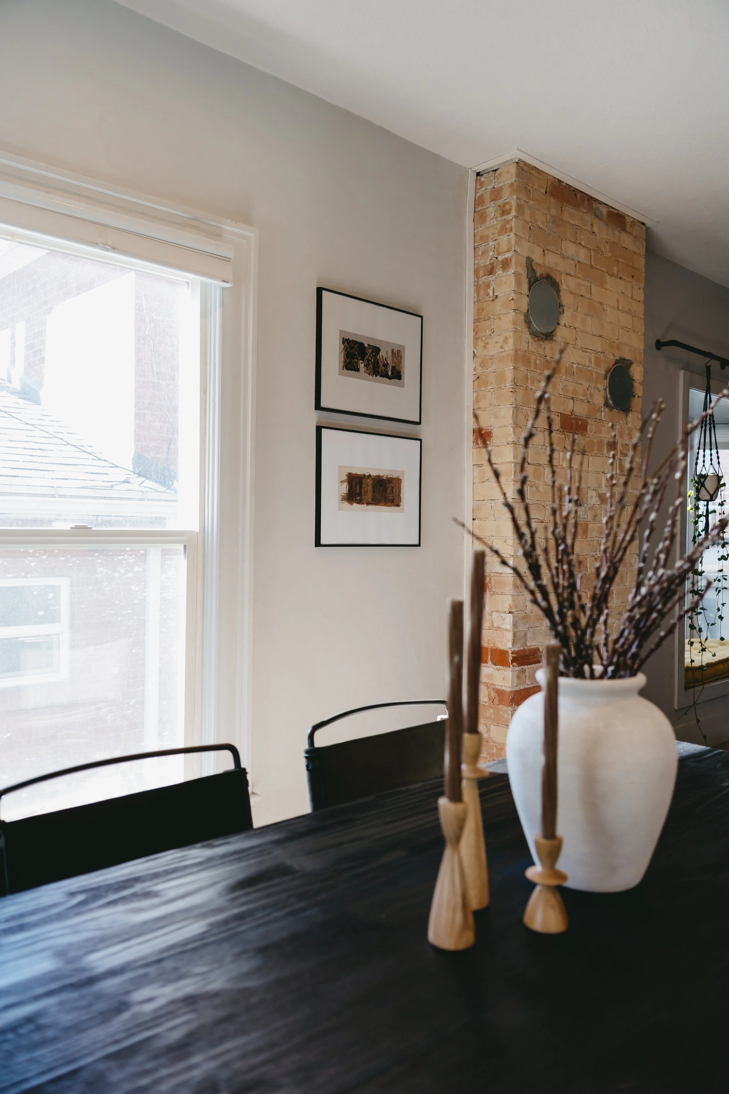 Riley James Interiors home staging and design. Interior of a dining room with a dark wooden table, a white vase with branches, and framed art on the wall, near a brick chimney and window letting in natural light.