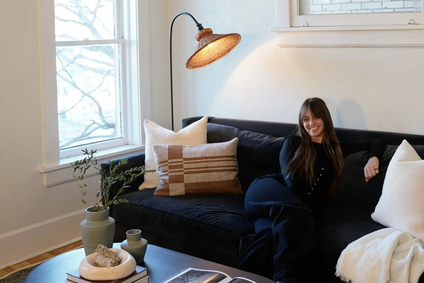 Woman with long hair smiling and sitting on a black sofa in a cozy living room with pillows and a white blanket, next to a window and a tall floor lamp with a wicker shade.