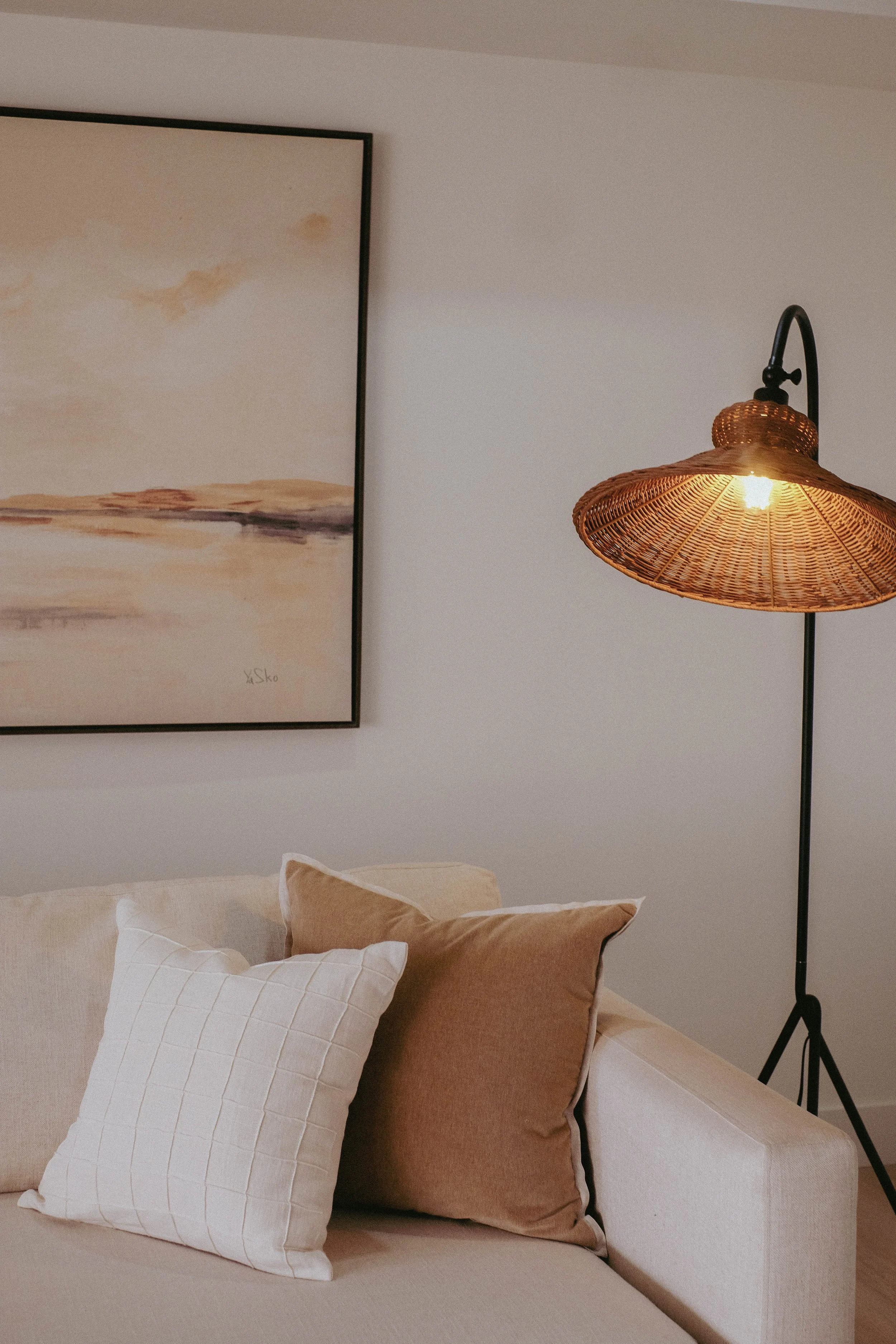 A bedroom with a beige bedspread, decorative pillows in white, beige, and pink, a framed abstract painting on the wall, and a wicker wall-mounted lamp with a black arm and stand.