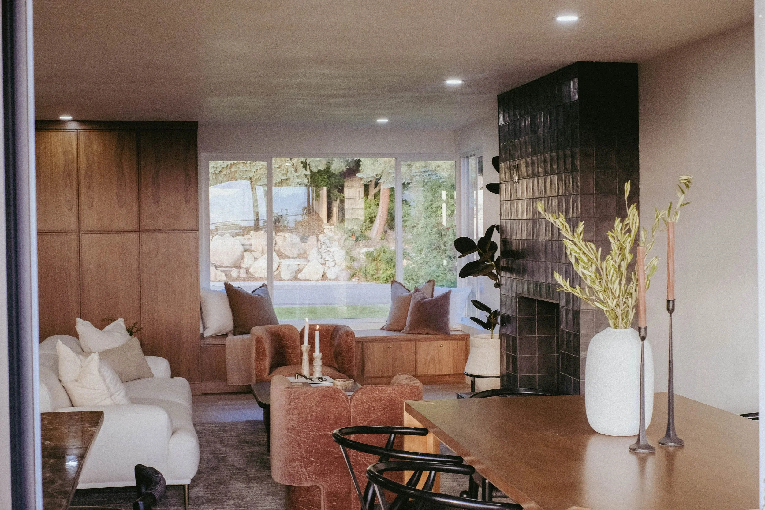 Living room with large window showing outdoor trees and rocks, beige and brown furniture, black tiled fireplace, and various decorative items.