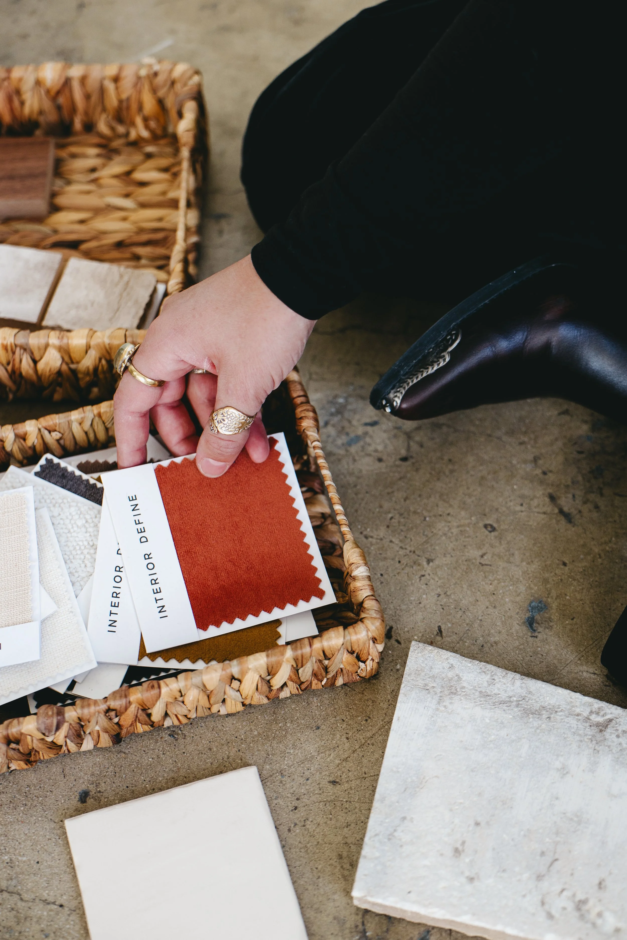 A person is browsing interior fabric samples from a basket, with tiles and fabric swatches on a concrete surface.