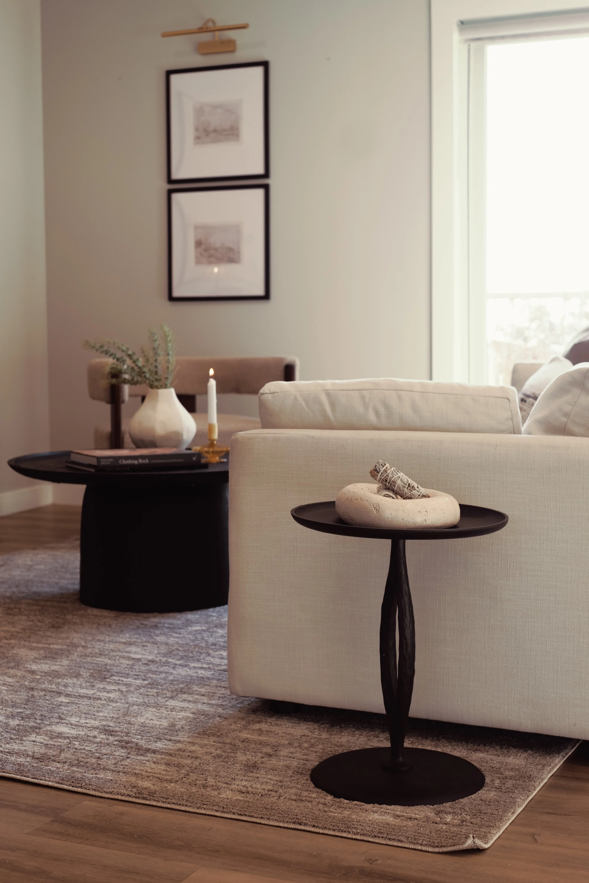 Living room with beige sofa, black side table with a white stone decor piece and rolled towels, soft textured rug, and large window with sunlight. Wall with two framed pictures and a decorative wooden art piece.