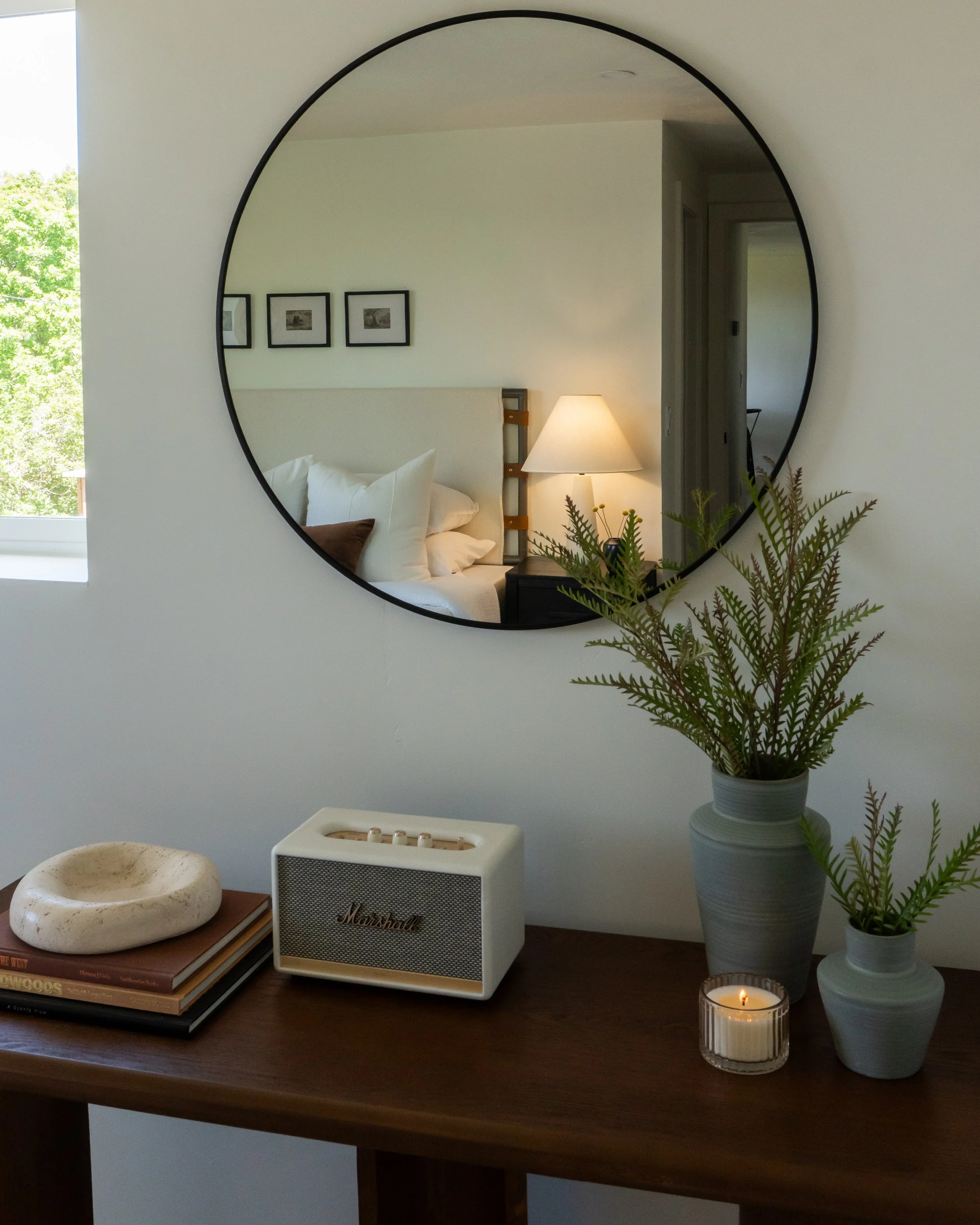 A circular mirror on a wall reflecting a bedroom with a bed, pillows, a nightstand with a lamp, and framed pictures. In front of the mirror, there is a table with a potted plant, a candle, a speaker, and some books.