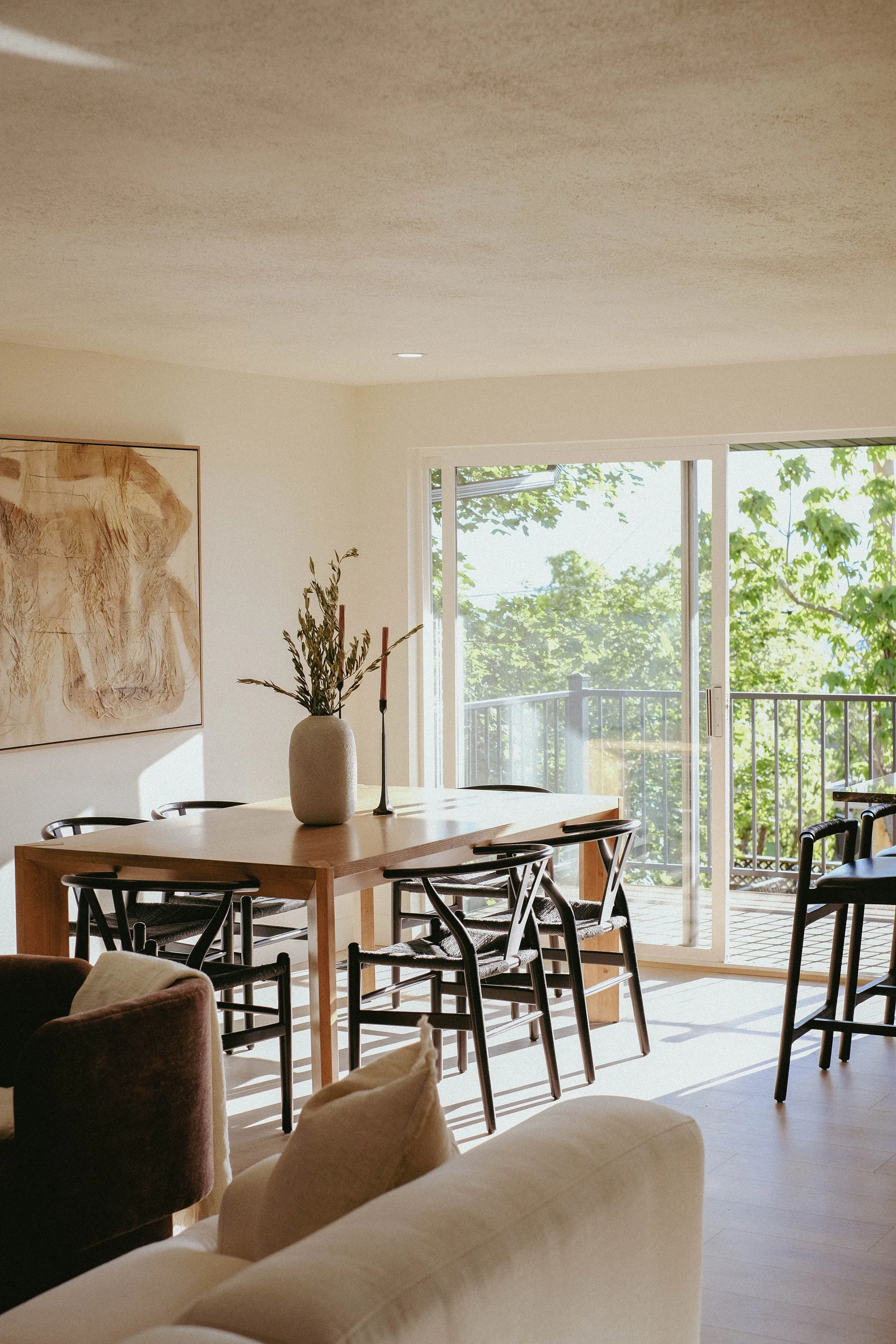 A dining area with a wooden table and black chairs, decorated with a large beige vase with green foliage, in a room with large windows showing green trees and blue sky outside.
