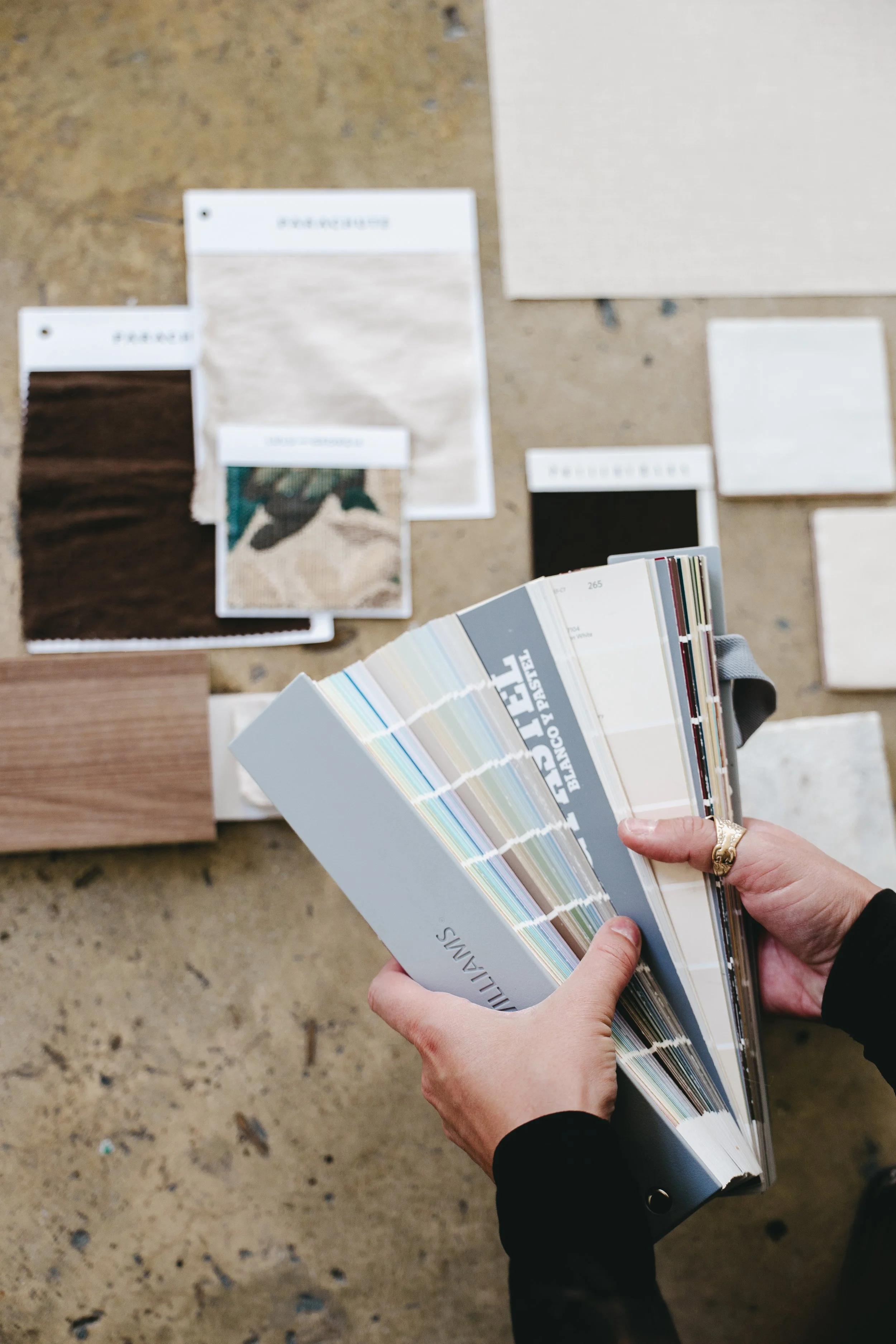 Person holding a color sample fan deck with various paint colors, surrounded by different sample boards and tiles on a concrete surface, used for interior design or remodeling.