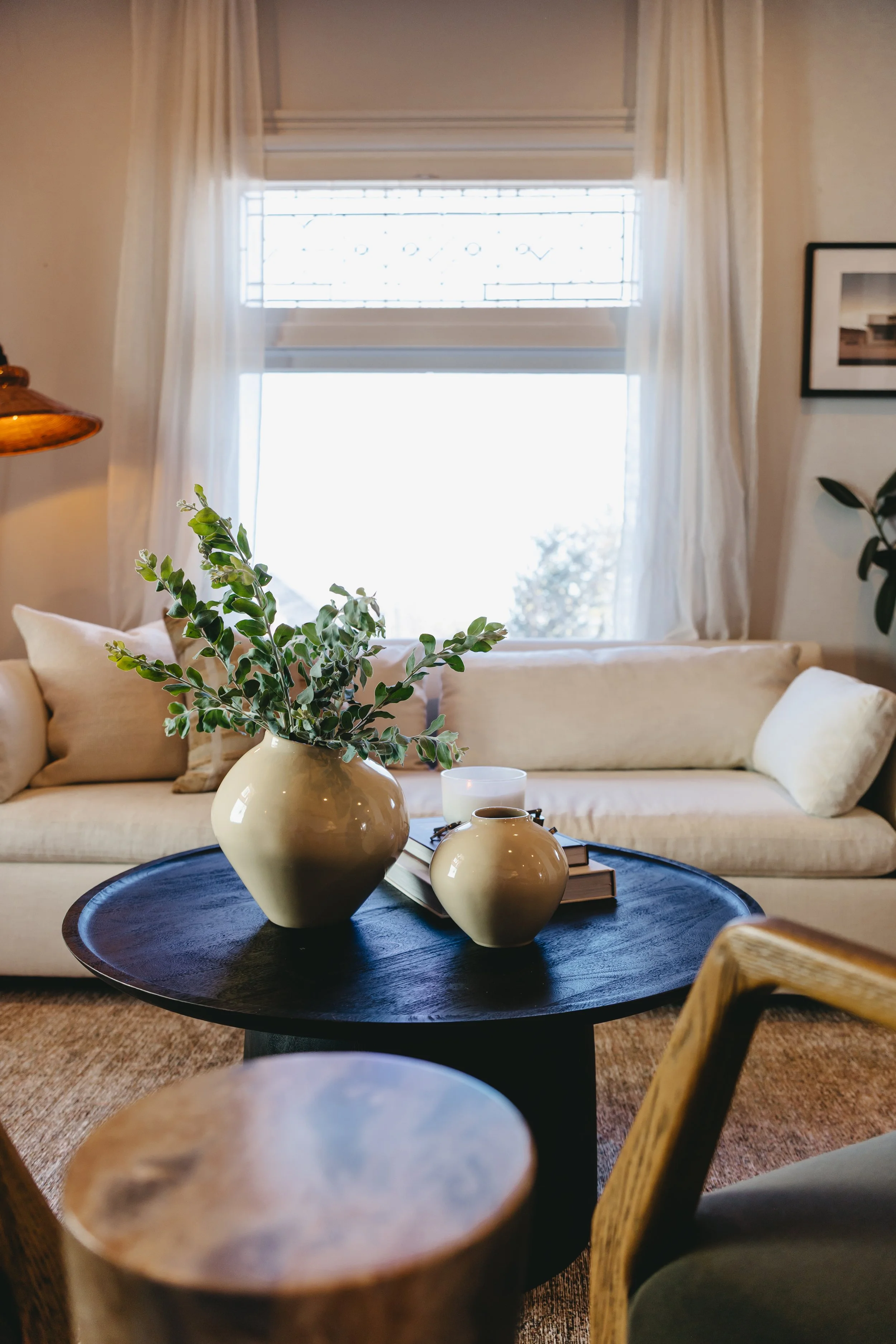 Vacant home staging by Riley James Interiors in Salt Lake City, Utah. Living room with a beige sofa, two beige vases with green foliage on a round black coffee table, and a window with white curtains.