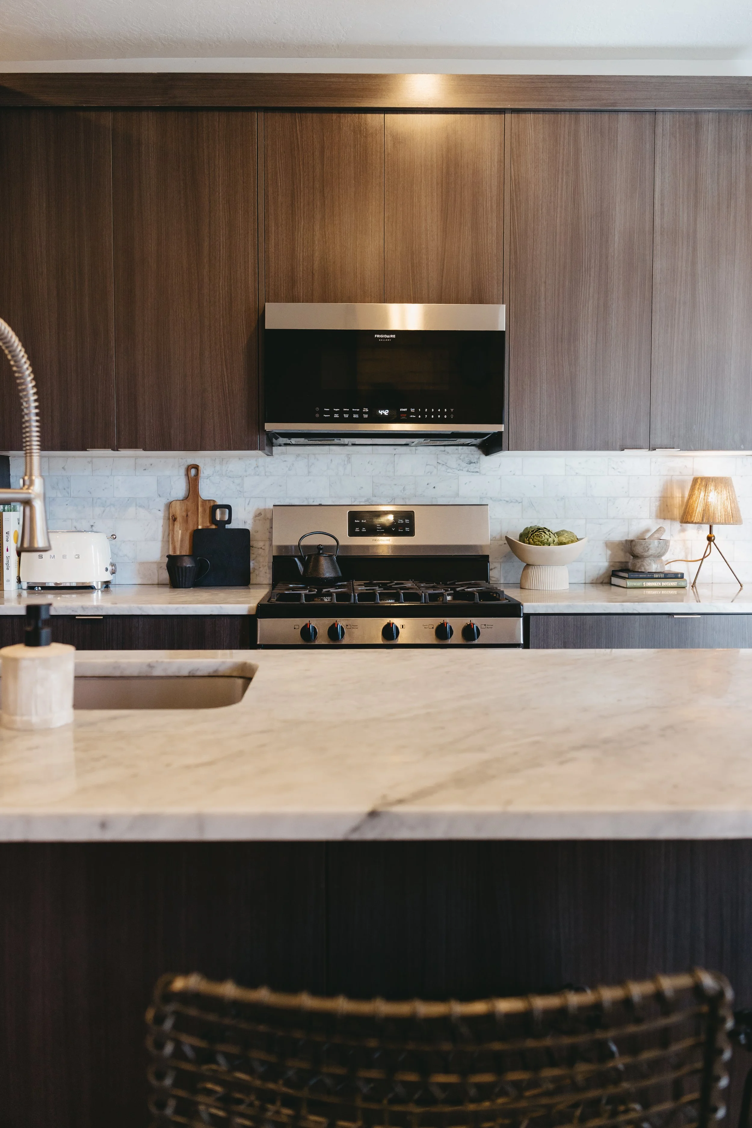 Modern kitchen with a marble countertop, wooden cabinets, stainless steel stove, black microwave, and decorative items including a bowl of artichokes and a small lamp. Vacant staging services by Riley James Interiors in Salt Lake City, Utah.