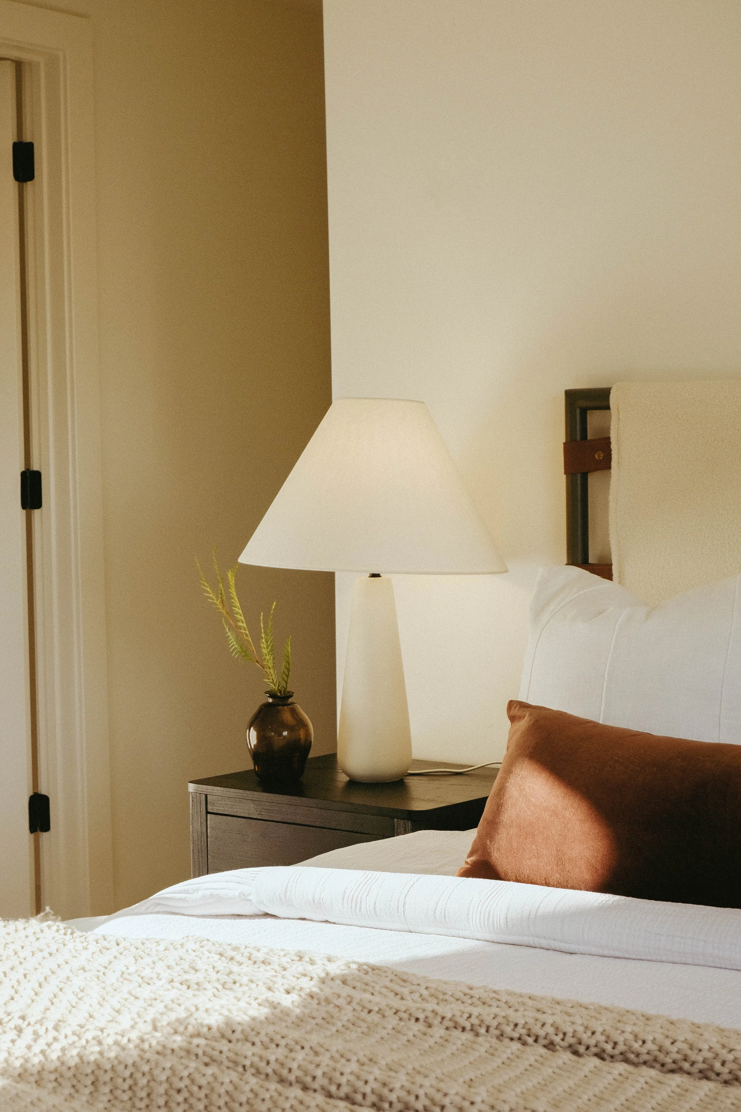 A bedroom with a bedside table holding a dark vase with a leafy green plant, a tall lamp, and a framed headboard. Part of the bed with a cream blanket and an orange pillow is visible.