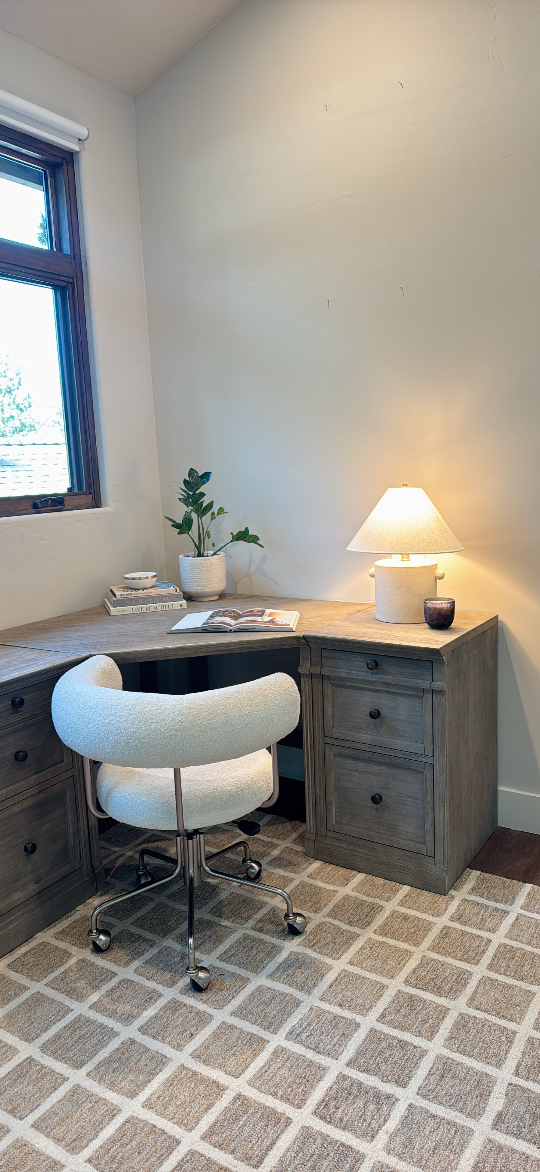 A cozy corner of a home office with a wooden desk, a white swivel chair, a table lamp, a small potted plant, an open book, and a window with a view outside. The room has beige carpet with a diamond pattern.