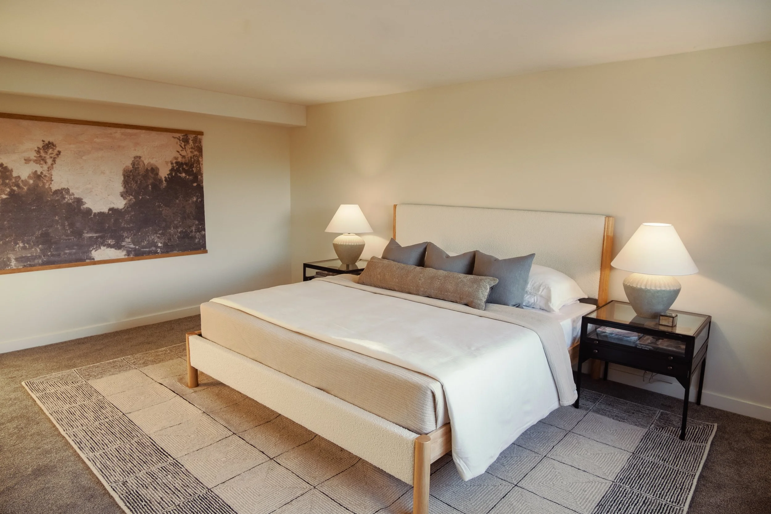 A bedroom featuring a large bed with a beige upholstered headboard, gray and beige pillows, and white bedding, illuminated by two bedside lamps on black nightstands, with a nature-themed painting on the wall and a patterned area rug on the carpeted f