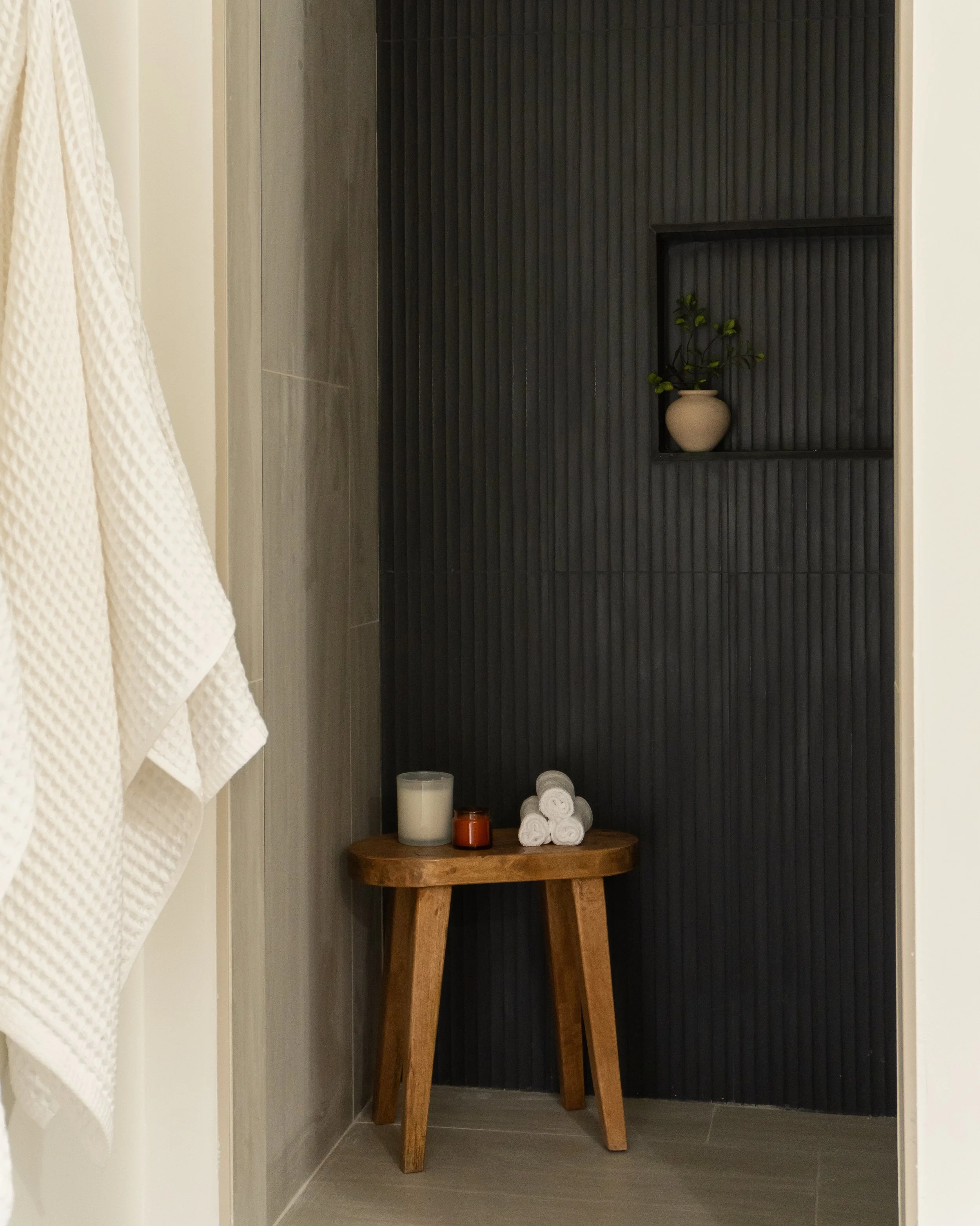 A small wooden table with rolled white towels, candles, and a potted plant against a black textured wall in a bathroom or spa setting.