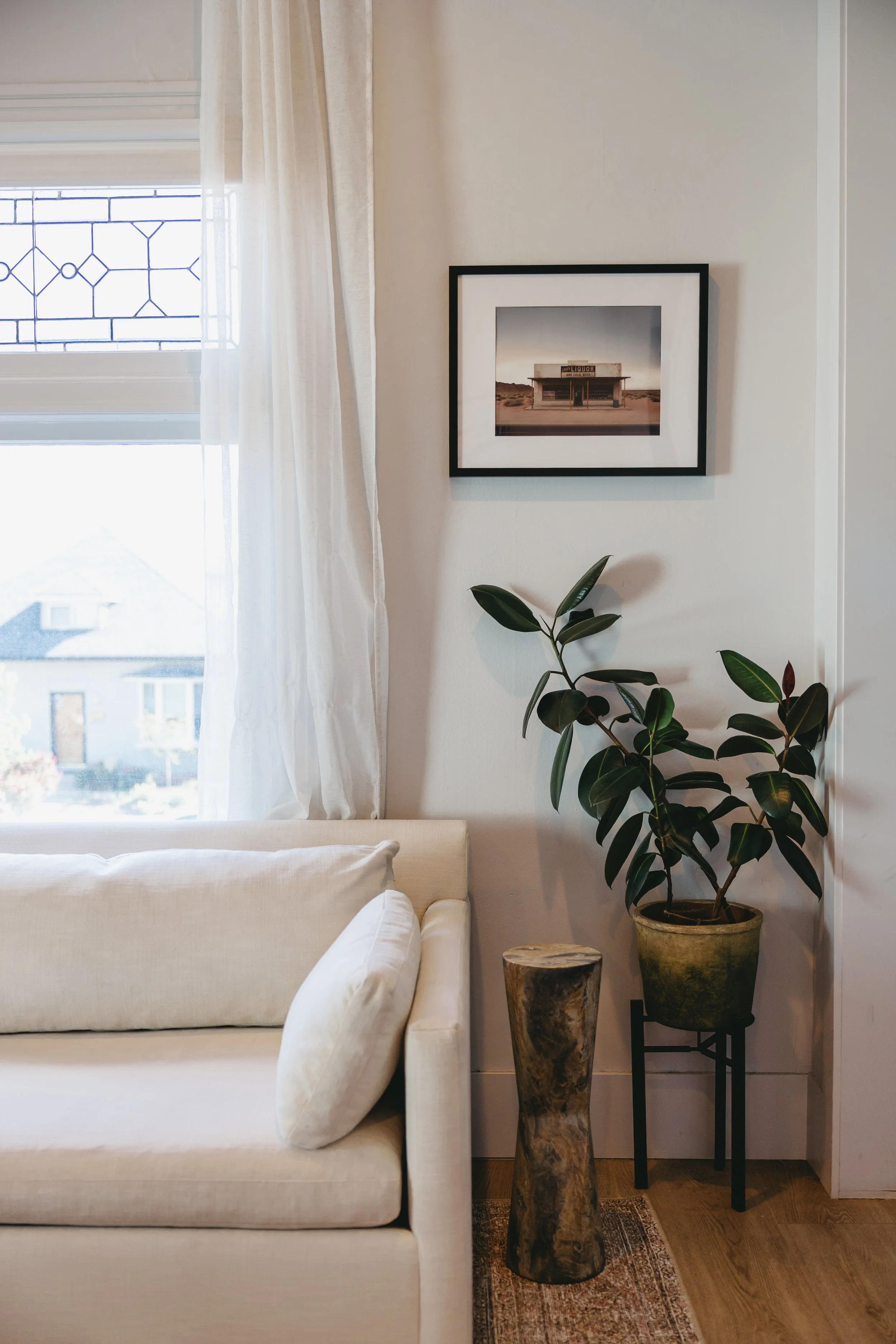 Interior view of a cozy living room corner with a white sofa, a large green plant in a pot on a stand, a wooden side table, framed picture on the wall, and a window with sheer curtains by Riley James Interiors in Salt Lake City, Utah.