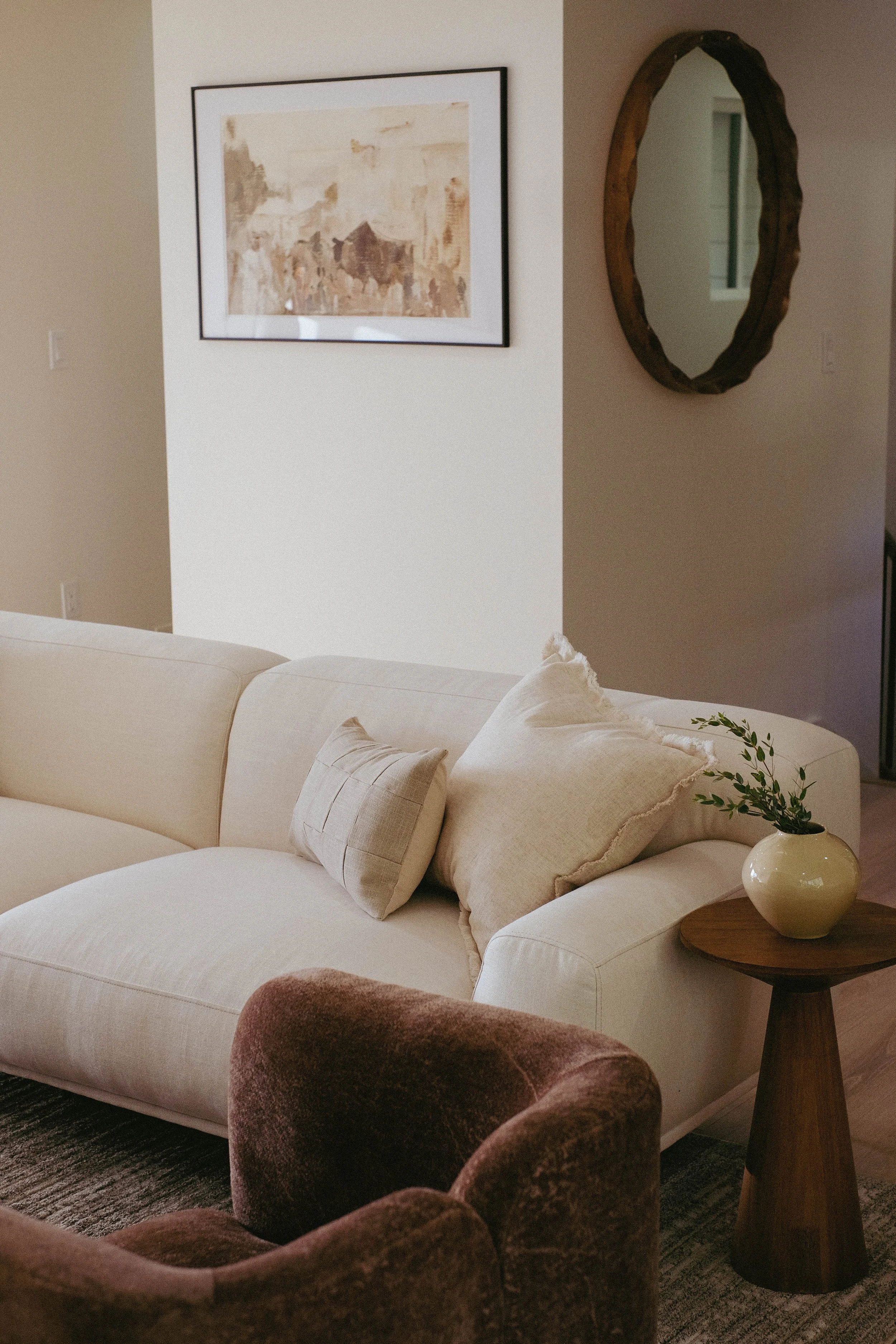 Living room with a white sofa, beige pillows, a brown velvet armchair, a small round wooden side table with a beige vase and plant, framed artwork, and a mirror on the wall.