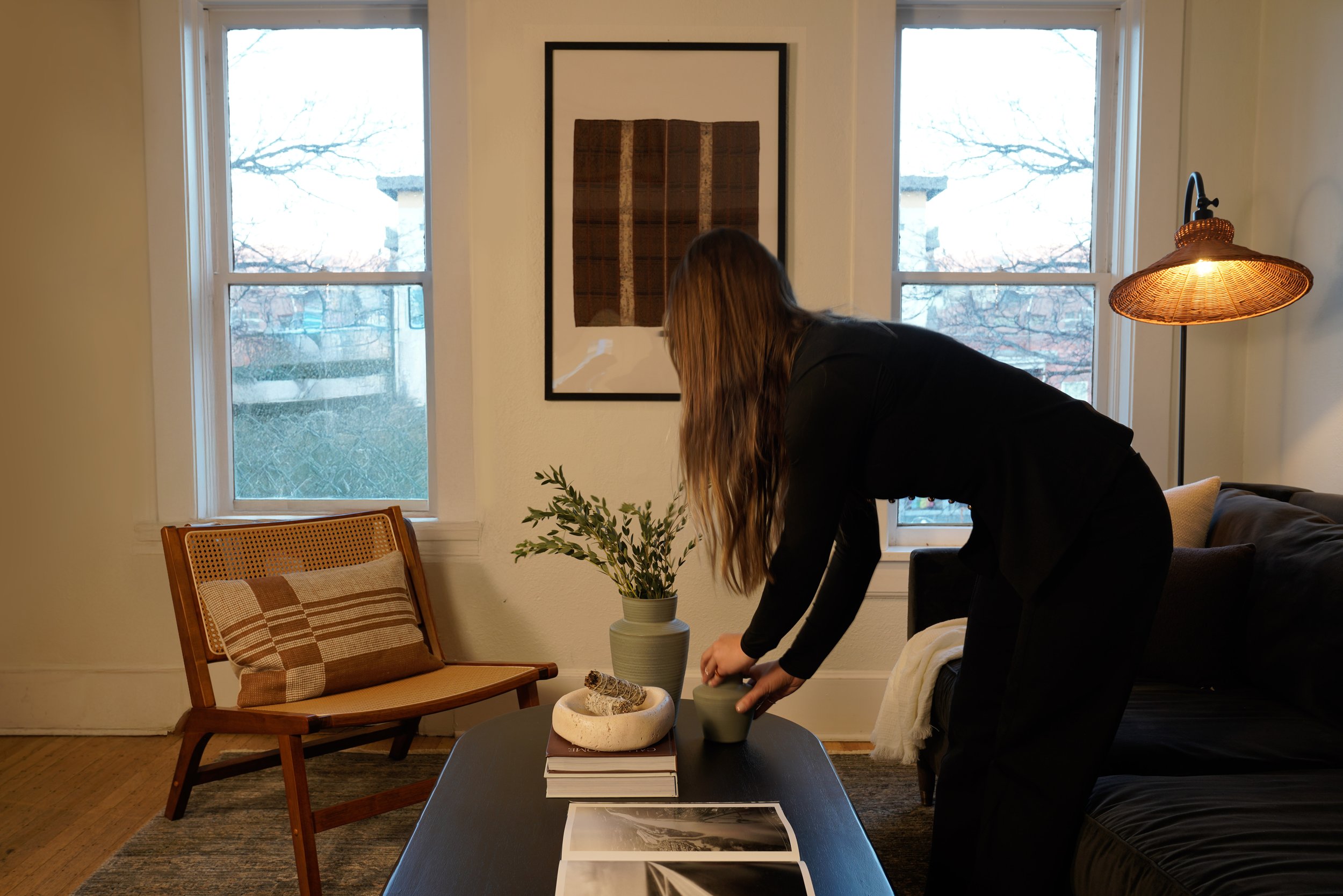 A woman with long hair arranging a potted plant on a dark coffee table in a living room with two large windows, a wooden chair, a framed artwork, and a sofa with cushions.