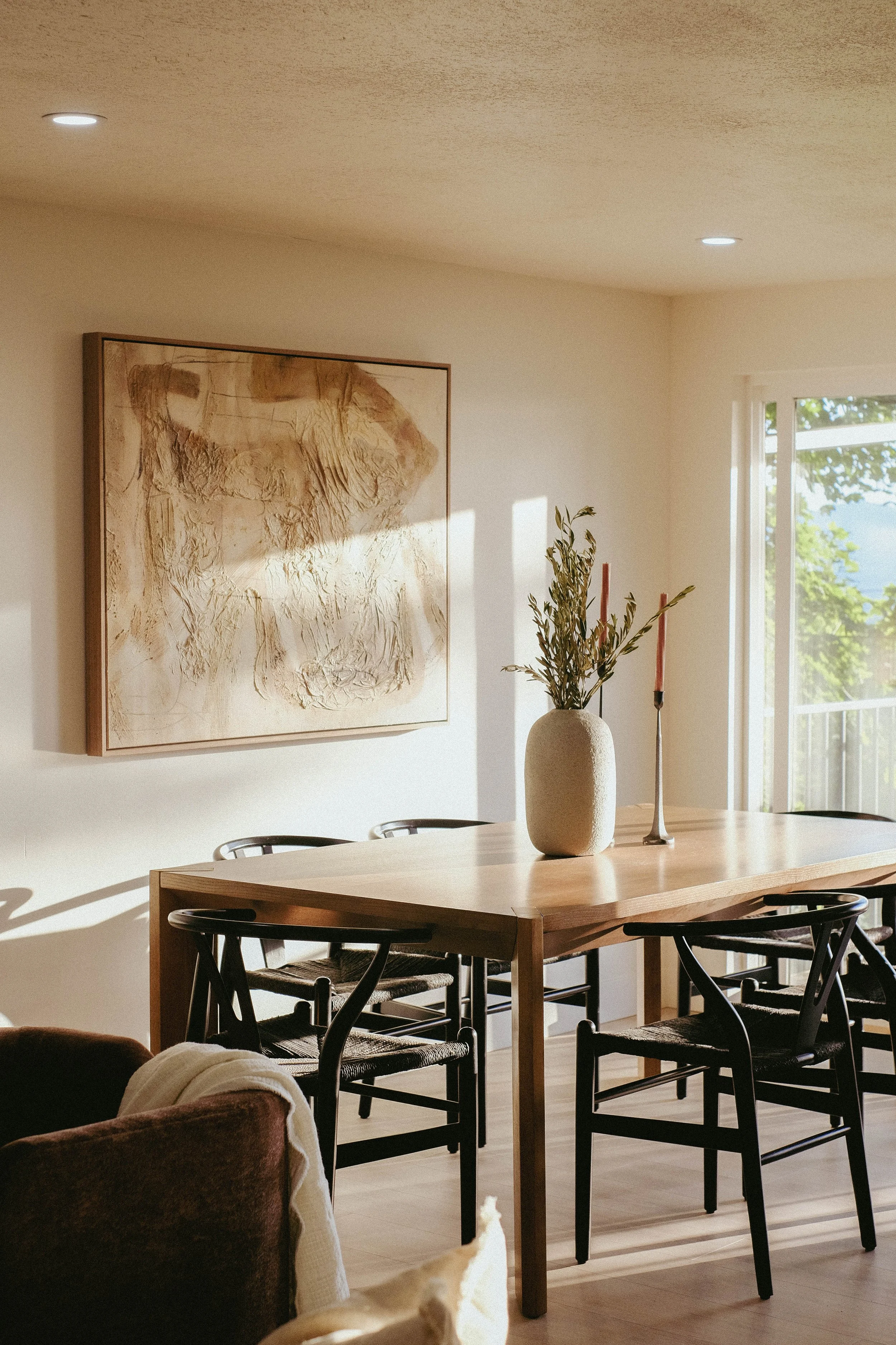 A dining room with a wooden table surrounded by black chairs, a large abstract art piece on the wall, a vase with green foliage, and pink candlesticks. Sunlight streams through a window showing greenery outside.