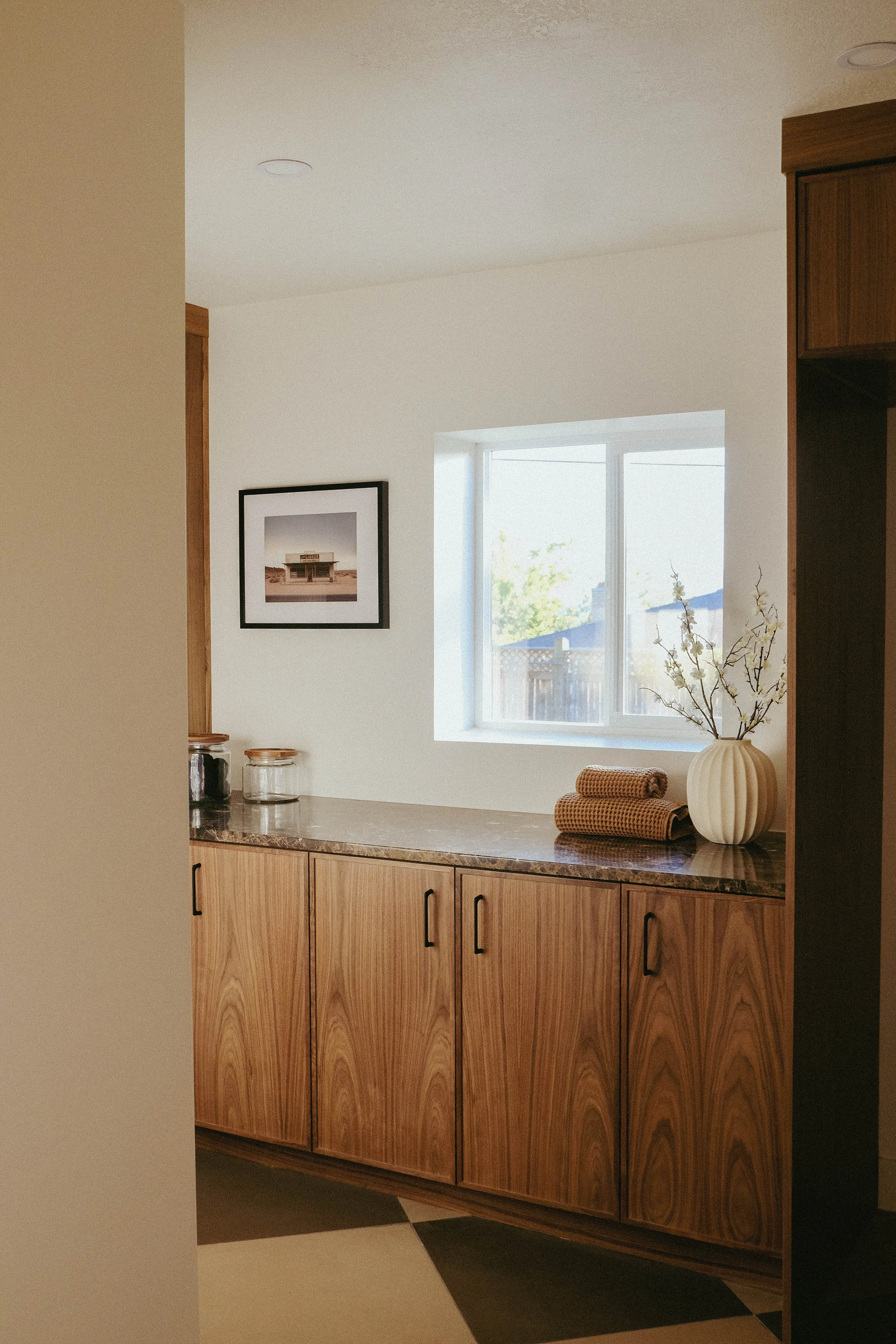 A room with a natural wood cabinet beneath a window, a beige vase with white flowers, and two brown towels, with a framed picture on the wall and jars on the countertop.