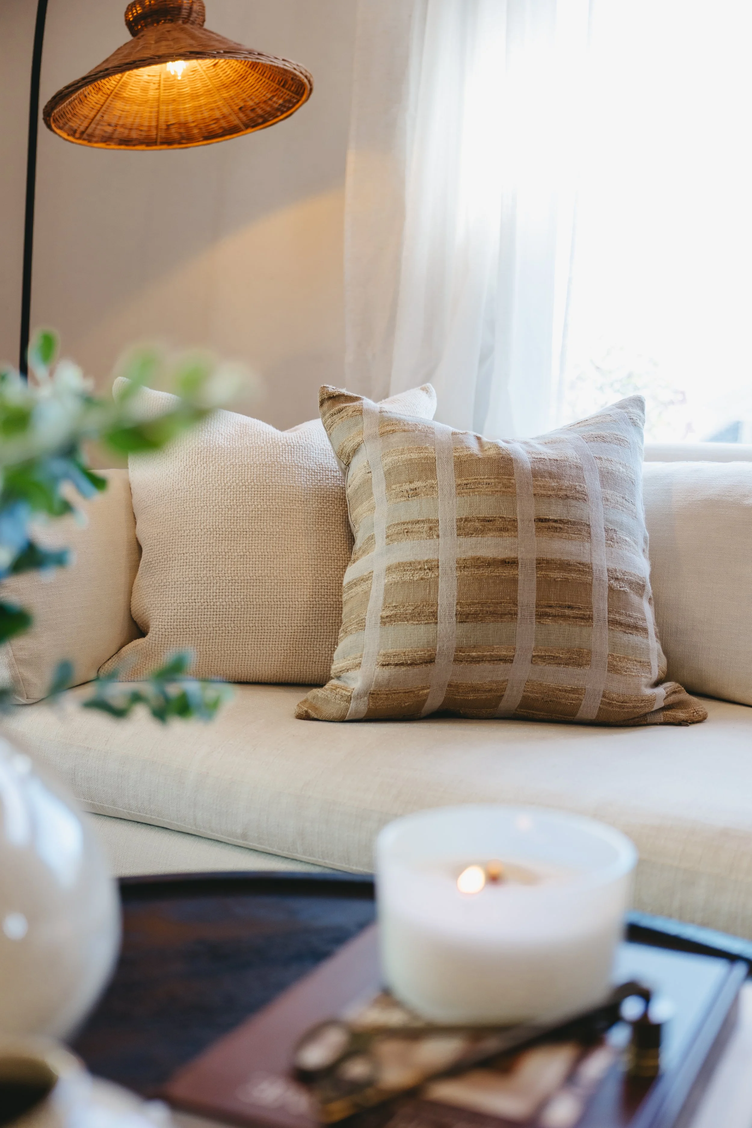 A cozy living room with a beige sofa and two decorative pillows, one with striped pattern and the other solid, near a window with sheer curtains. Staging by Riley James Interiors in Salt Lake City, Utah.