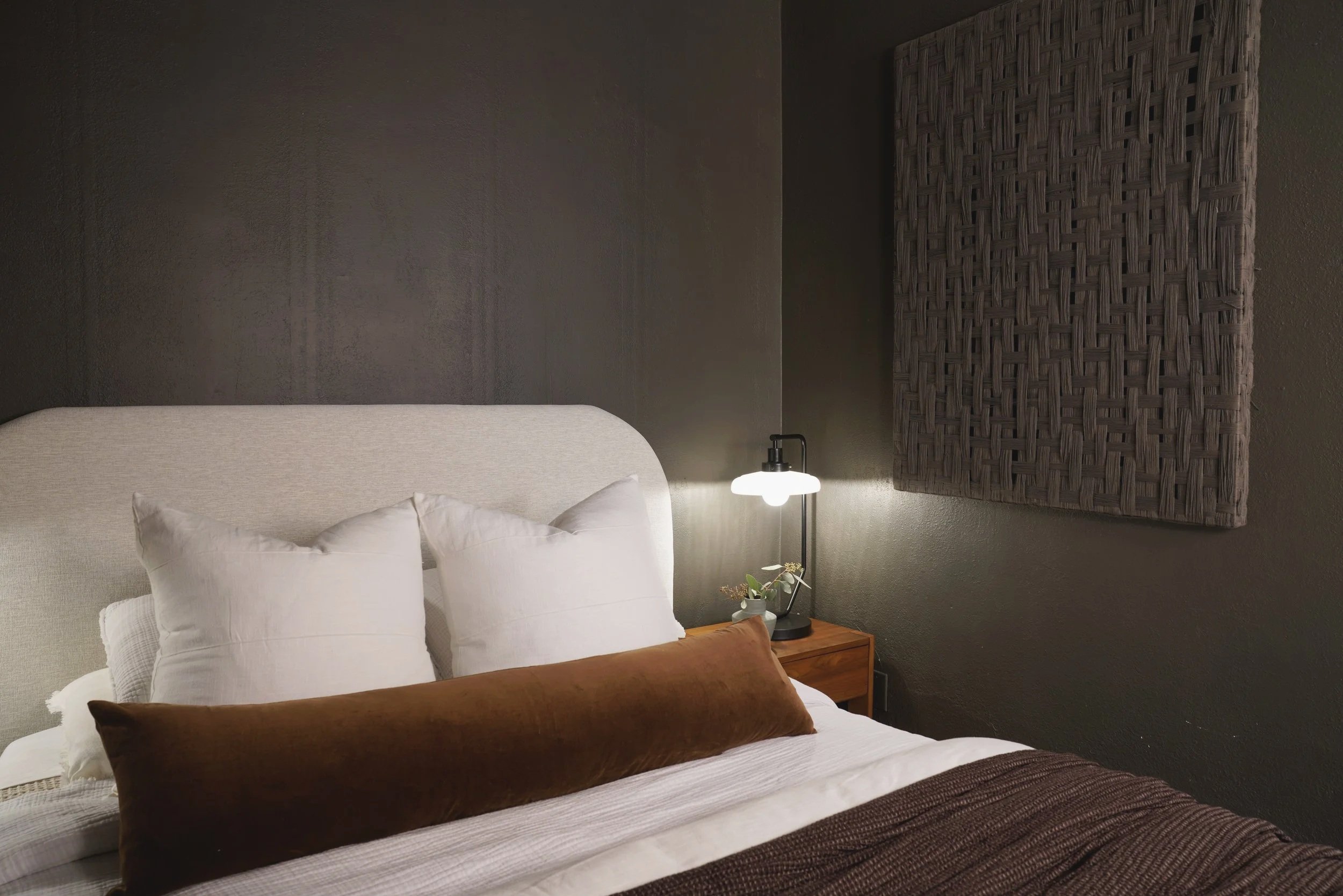 A cozy bedroom with a beige upholstered headboard, white pillows, a brown cushion, and a textured wall art piece. A small wooden bedside table with a black lamp and a small potted plant. The wall behind is a dark, textured wall.